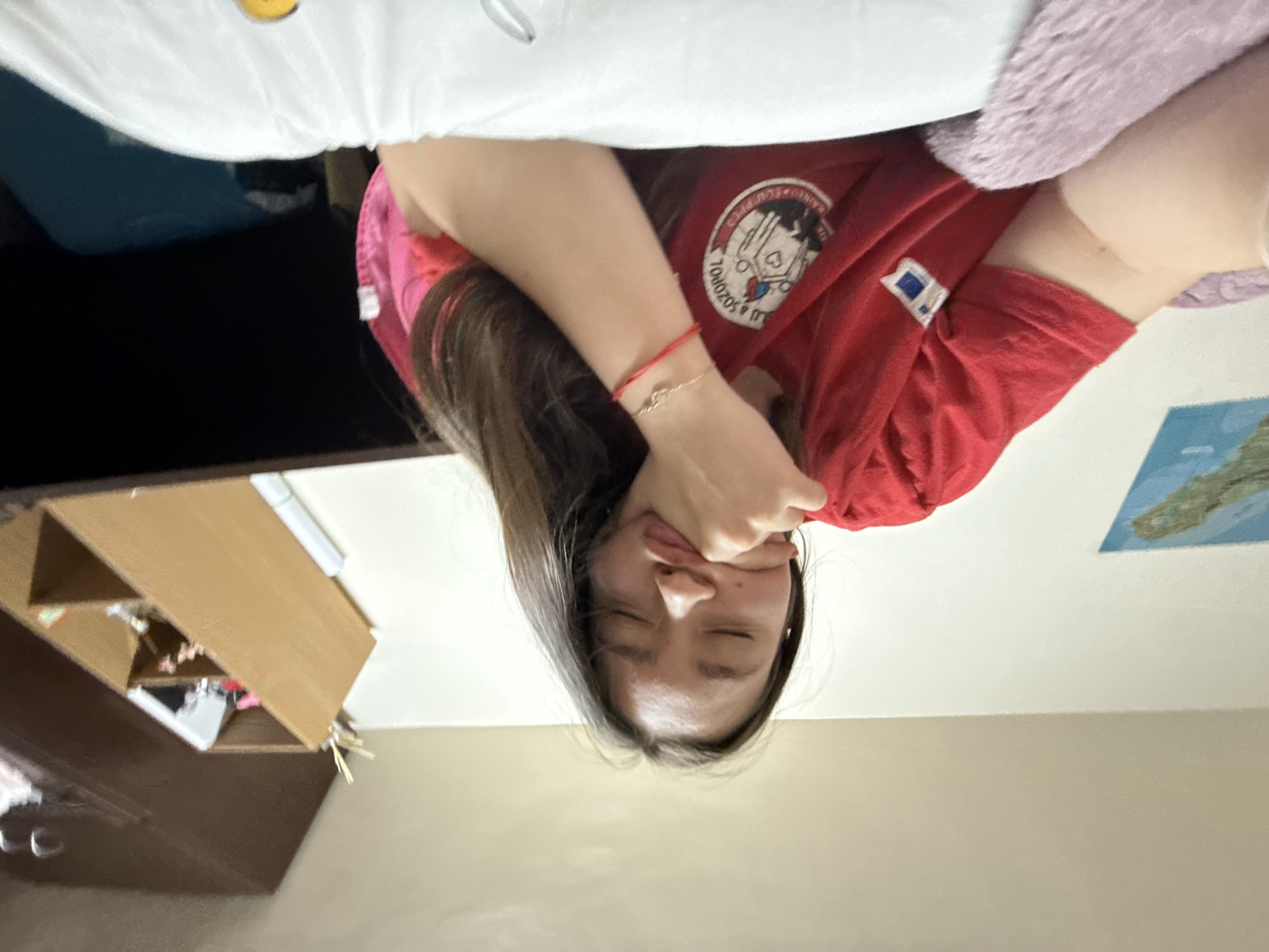 A young woman is upside down in what appears to be a bedroom. She is wearing a red t-shirt and holding her cheeks with her hands, causing her face to be squished. The room contains a bed with white sheets, a fuzzy pink blanket, and a dark-colored dresser with a shelf above it. A map is visible on the wall. It is not possible to determine the location in Sofia, Bulgaria from this image.