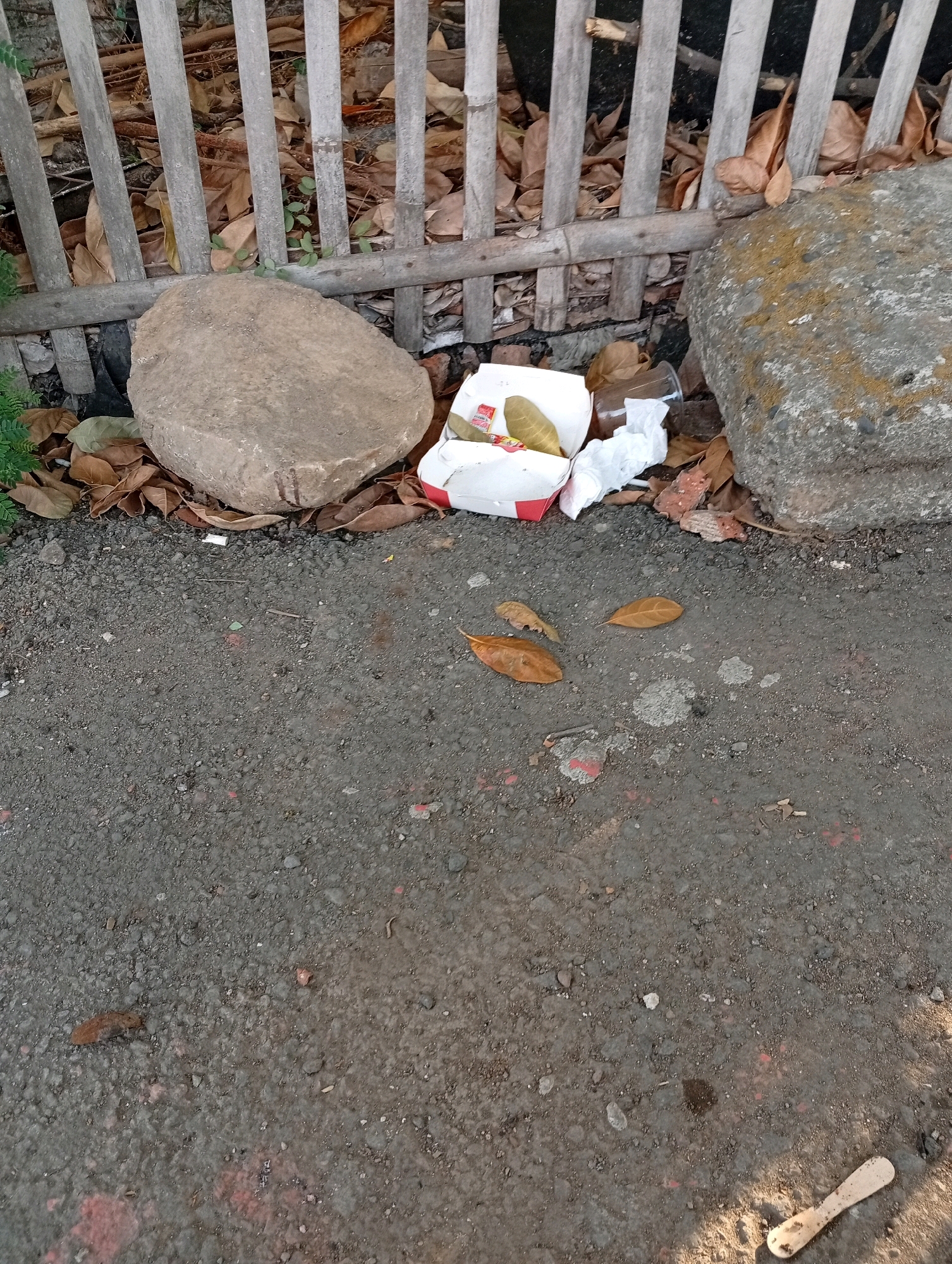 The image depicts an outdoor scene near a bamboo fence with dry leaves scattered around. Two large rocks are present, one on the left and one on the right. In the center of the image, a discarded fast-food container with a white and red design sits on the ground, containing what appears to be two pieces of food. Crumpled white paper and a clear plastic cup are also discarded near the container. The ground is made of asphalt with small pebbles. Several dry leaves are scattered on the asphalt, and some green foliage is visible near the bamboo fence. The scene appears to be in a public area, possibly a roadside or park, in Karawang, Indonesia.