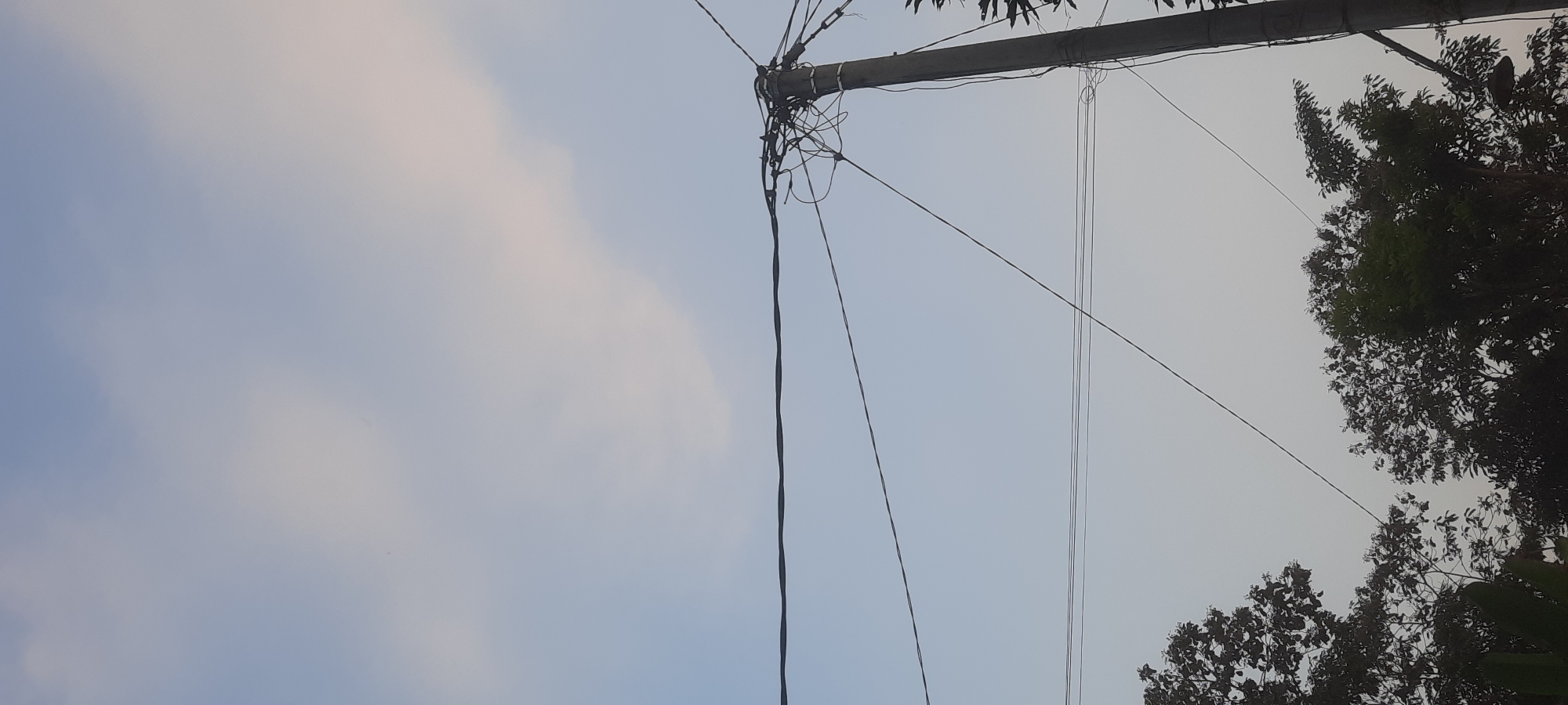 The image depicts a utility pole with numerous tangled wires against a pale blue sky with wispy clouds. The pole, made of concrete, is positioned on the right side of the frame. Several horizontal and diagonal wires are strung across the sky, some connected to the pole and others running independently. At the bottom of the image, the dark silhouettes of trees are visible, suggesting an outdoor setting, possibly in a rural or suburban area. The lighting indicates it is daytime, but the sky's paleness suggests it might be early morning or late afternoon. The location is Ploso, Indonesia.