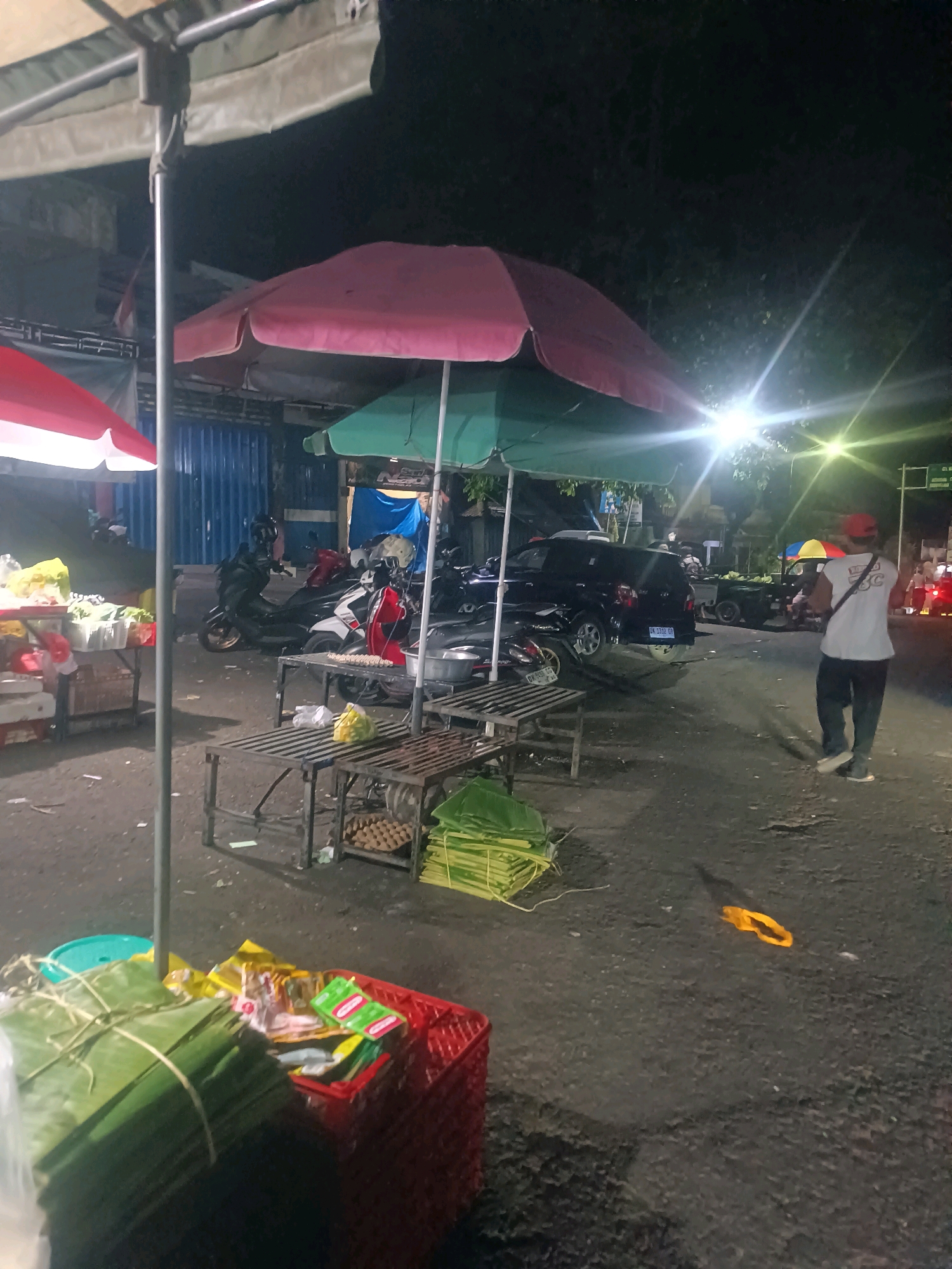The image captures a nighttime scene at a market stall in Tabanan, Indonesia. A person wearing a white t-shirt, dark pants, and a red cap stands facing away from the camera, walking on an asphalt surface. They have a bag slung over their shoulder. Several market stalls are visible, covered by colorful umbrellas—pink, green, and red. In the background, motorcycles are parked, and a dark-colored SUV with a license plate that reads "DK 1312 GR" is present. A yellow object, possibly a piece of clothing or material, lies on the ground. The ground is littered with debris, and a bright light source illuminates the area from the upper right. Stacks of green leaves, presumably for packaging or food preparation, are organized on tables. Various items are displayed on the tables, including what appear to be food products and egg cartons. The overall impression is that of a lively, albeit slightly cluttered, night market environment.