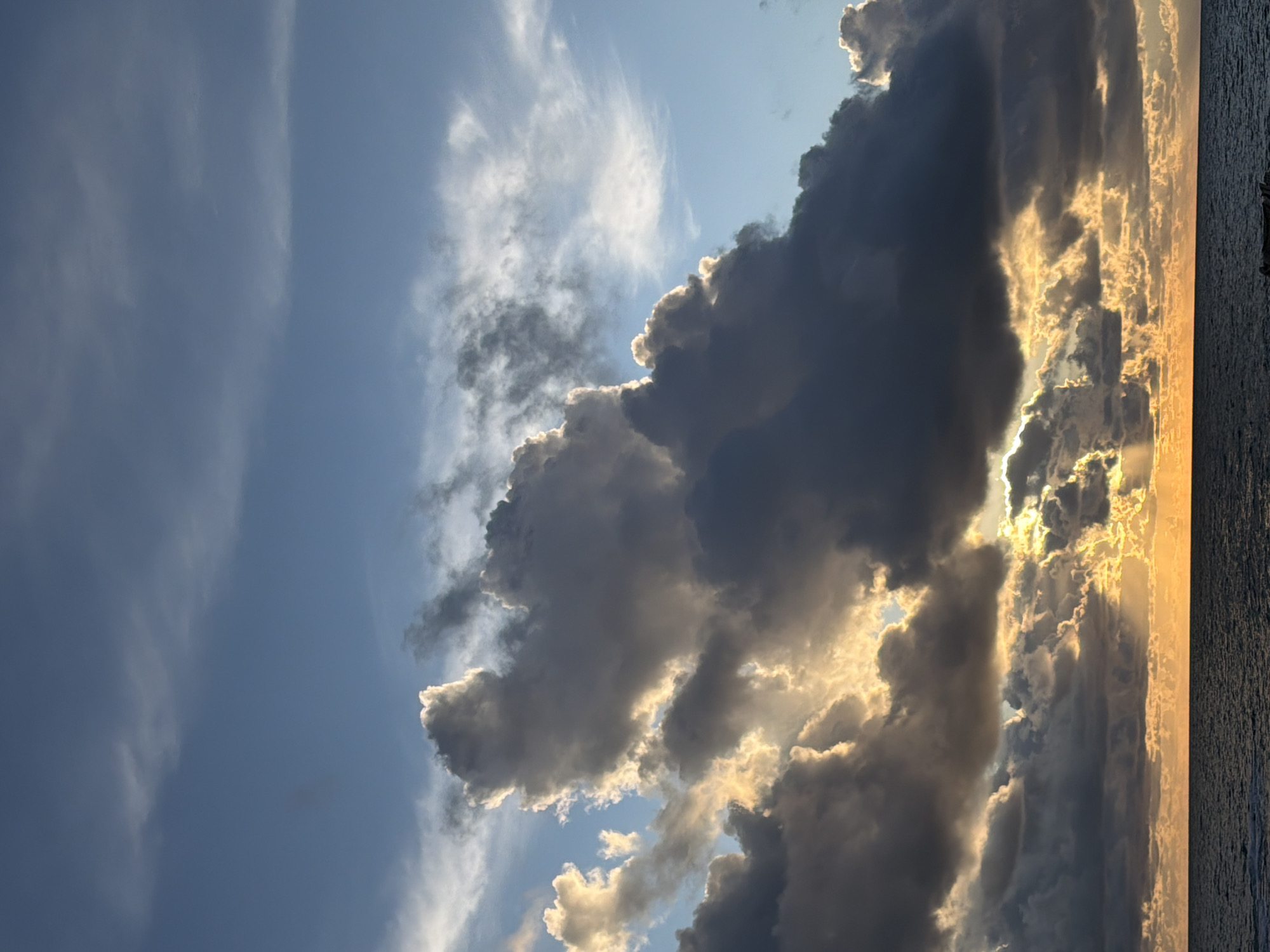 The image depicts a seascape at sunset, observed from the coastline of Tel-Aviv, Israel. The upper two-thirds of the frame consist of a sky transitioning from a light blue at the top to a dramatic configuration of clouds below. Large, dark grey cumulus-type clouds dominate the mid-ground, heavily backlit by the setting sun, which is obscured from direct view. Intense golden-orange light emanates from behind and through these clouds, illuminating their edges and creating distinct crepuscular rays that extend towards the horizon. The lower third of the image shows a dark blue-grey body of water, identified as the Mediterranean Sea, exhibiting minor surface ripples. A prominent, unbroken horizontal line marks the horizon, where a concentrated band of golden-orange light is present. No persons, vessels, or man-made structures are visible within the frame.