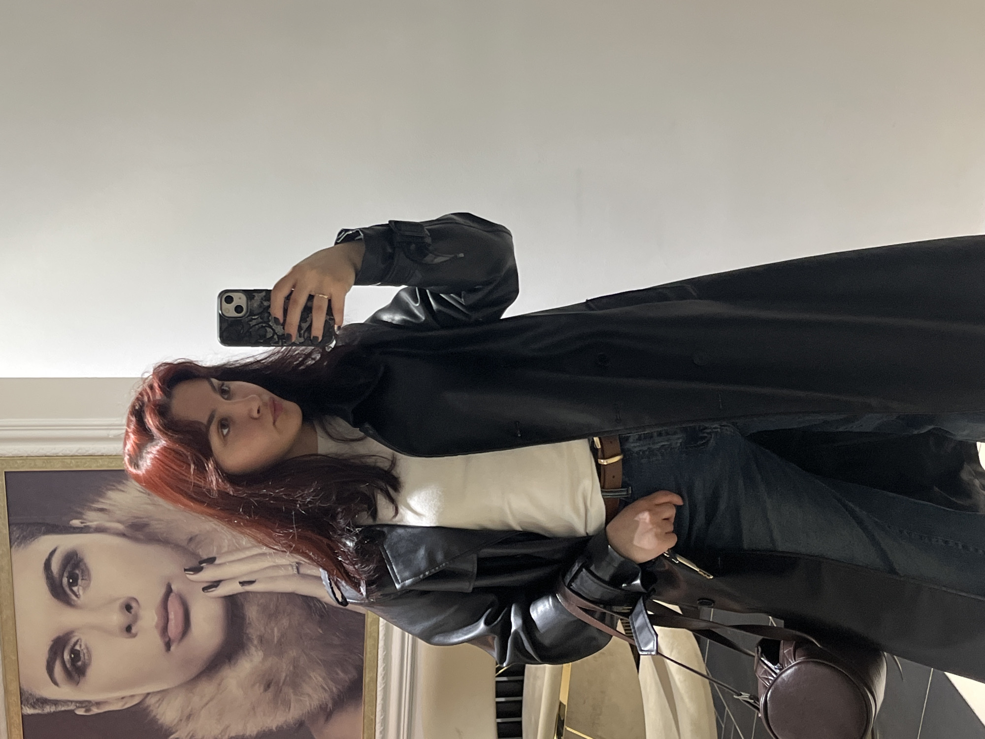 A woman with red hair takes a selfie in front of a framed portrait of another woman. She is wearing a white shirt, blue jeans with a brown belt, and a long black leather coat. She holds a cell phone in her left hand and a set of keys in her right. A brown leather purse hangs from her right shoulder. The background is a light-colored wall. There is no information in the image to indicate it was taken in Neftekamsk, Russia.