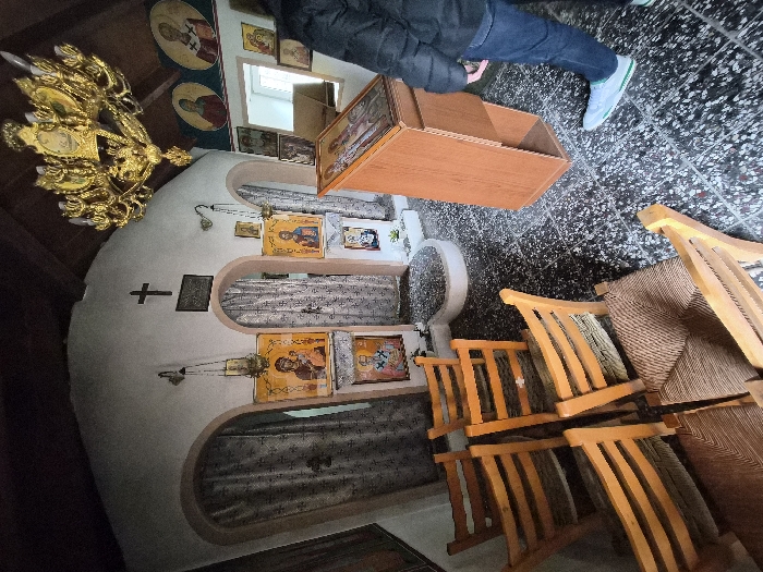 The interior of a small Orthodox chapel in the Municipal Unit of Loutraki - Perachora, Greece, is shown from a low angle. The room features light-colored, textured walls and a dark wooden ceiling. The floor is covered with dark, speckled square tiles. A black Latin cross is mounted on the central wall, positioned above three arched recesses or doorways draped with light-colored, patterned fabric curtains. Multiple religious icons are displayed on the walls, including several framed rectangular icons at eye level and circular icons positioned higher on the right wall. Two brass oil lamps hang from chains in front of icons. A large, ornate golden chandelier with multiple light fixtures is suspended from the ceiling on the right. In the foreground-left, six light-brown wooden chairs with woven rush seats are arranged in rows, facing away from the viewer. To the right, a wooden lectern with an icon displayed on its slanted top stands upright. A person is partially visible on the far right, from the waist down, wearing dark blue jeans and light blue/grey athletic shoes with green accents on the soles. The person is standing, oriented towards the lectern and the icons on the wall.