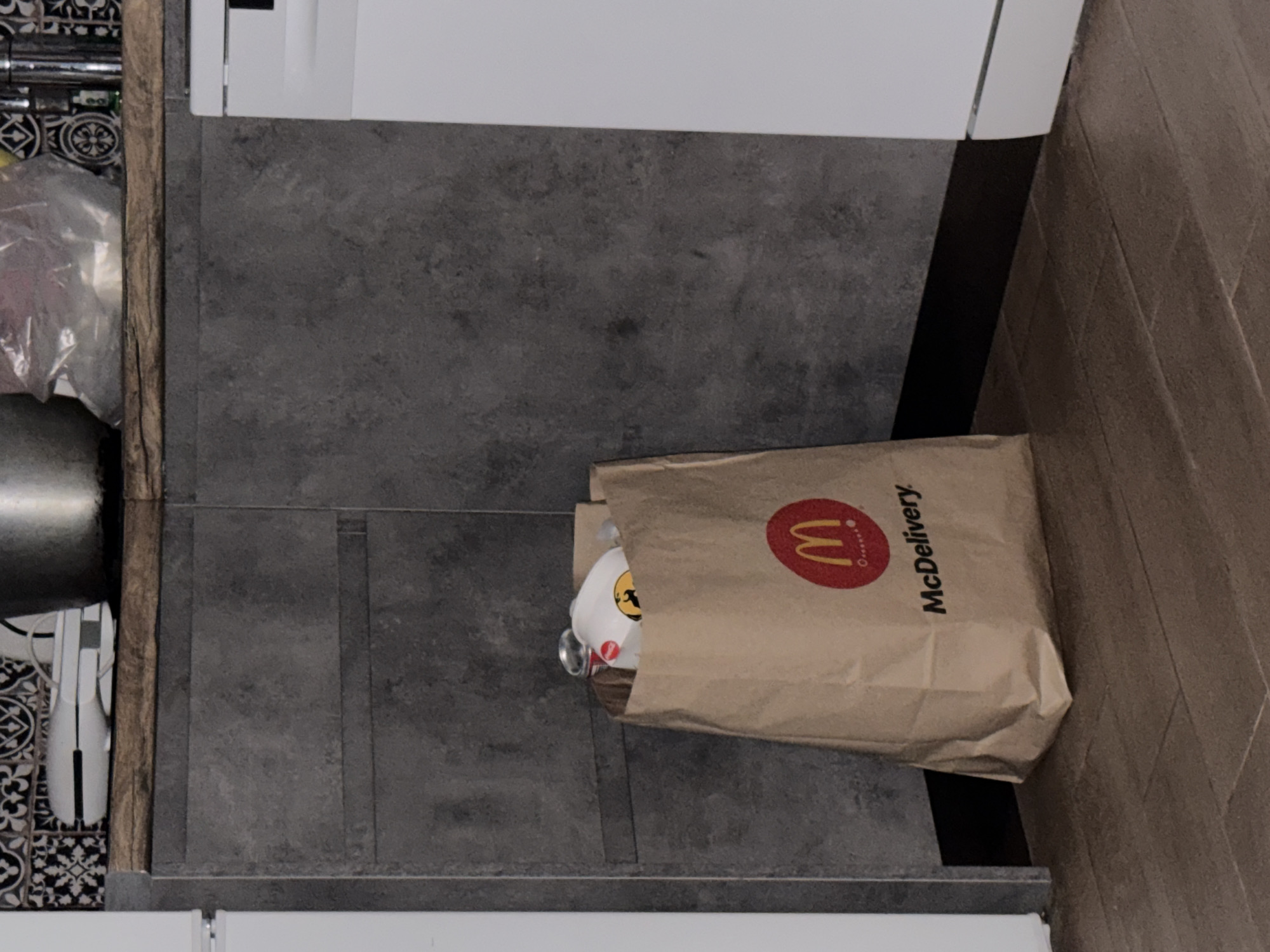 In an indoor setting, likely a kitchen, a brown paper McDonald's McDelivery bag stands on a wooden floor. The bag is partially filled with items, one of which appears to be a white paper cup with a red logo. A silver can is also visible within the bag. Behind the bag, a dark grey tiled wall forms the backdrop. To the left of the bag, a white appliance, possibly a dishwasher or refrigerator, is partially visible. To the right, another white appliance is also present. The upper portion of the image reveals a countertop with some objects, including what appears to be a large metal bowl and a white appliance. The overall scene suggests a casual, domestic environment.
