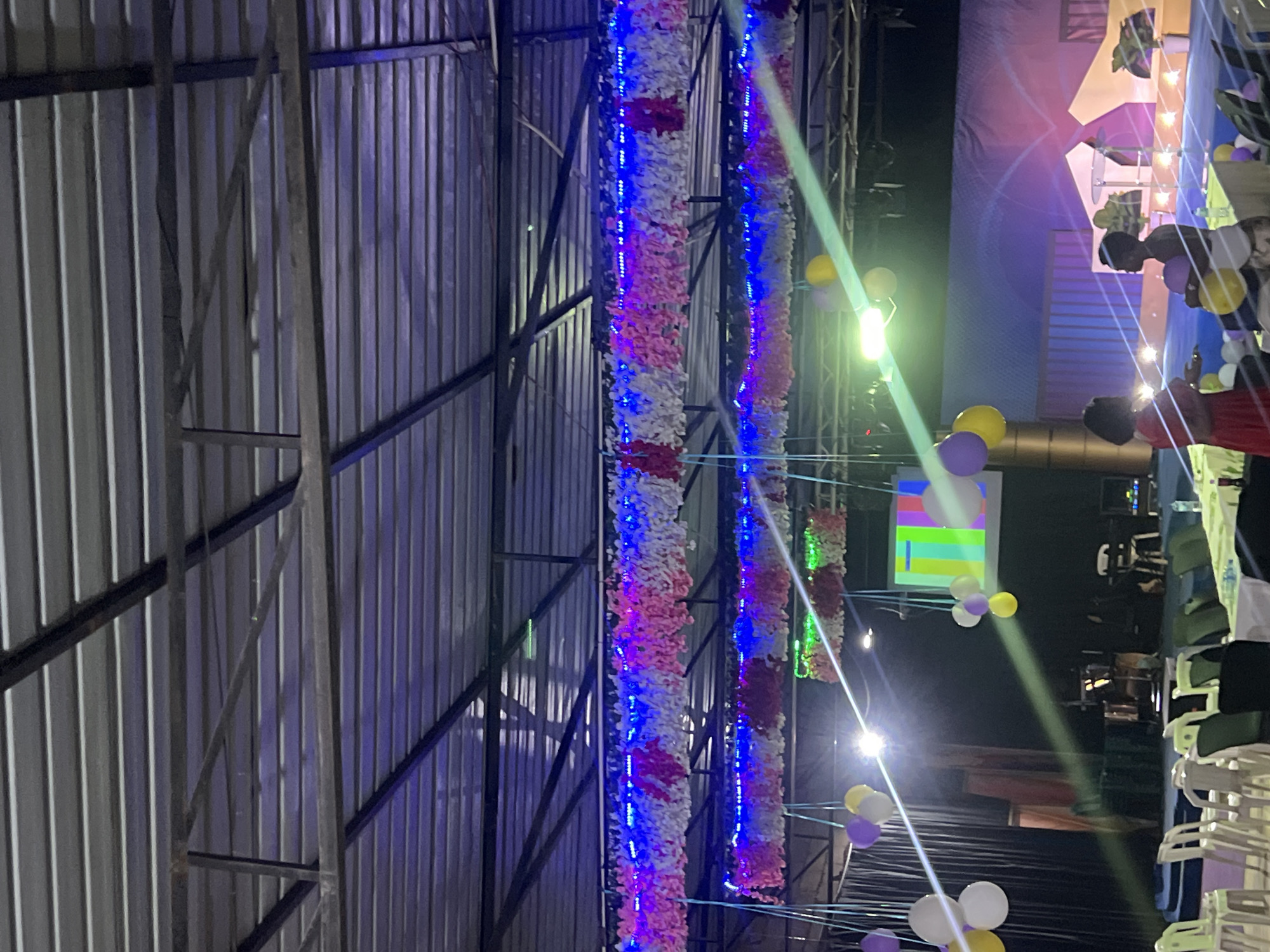 The image depicts an indoor event space, likely a hall or covered outdoor venue, in Durumi, Nigeria. The ceiling is constructed with corrugated metal sheeting supported by a metal framework. Festive decorations hang from the ceiling, including strings of pink, white, and blue artificial flowers interwoven with blue LED lights. Balloons in white, yellow, and purple are also suspended, some in clusters and others individually.
In the background, a screen displays a colorful static pattern resembling a test pattern. Stage lighting casts diagonal beams across the scene. The seating area consists of rows of white plastic chairs and some darker chairs, with tables covered in white cloths. A few individuals are visible. In the foreground, two women are standing, one in a red dress and the other in a white top and patterned skirt, appearing to be engaged in conversation. In the middle ground, to the right, a man in a white shirt and dark shorts is holding balloons. Further back, musical instruments, including drums and what appears to be a keyboard, are set up on a stage area. The overall atmosphere suggests a celebratory event, possibly a wedding reception, party, or community gathering.