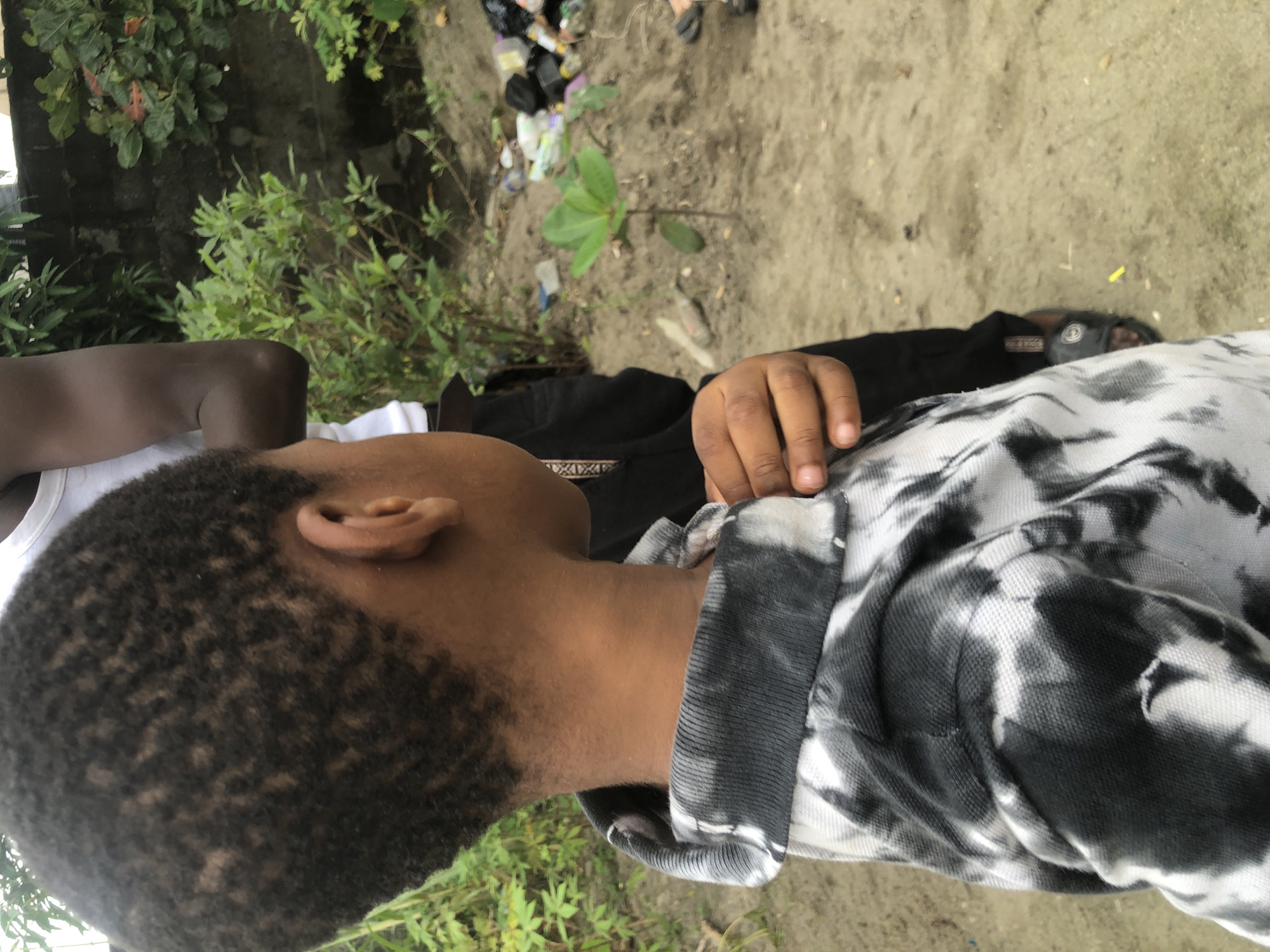 The image depicts a close-up of a young boy from the shoulders up, facing away from the camera and looking to his right. He has dark, textured hair and is wearing a black and white tie-dye polo shirt. His right hand is visible, touching the collar of his shirt. Behind him, another person, whose face is not visible, is partially in frame, wearing a white tank top and black pants. The environment appears to be outdoors, with a sandy ground and dense green foliage in the background. To the right, a wall is visible, with some debris, including plastic bottles and bags, scattered on the ground. The scene is consistent with an outdoor setting in a less developed area, possibly a backyard or undeveloped plot of land within Ado, Nigeria.