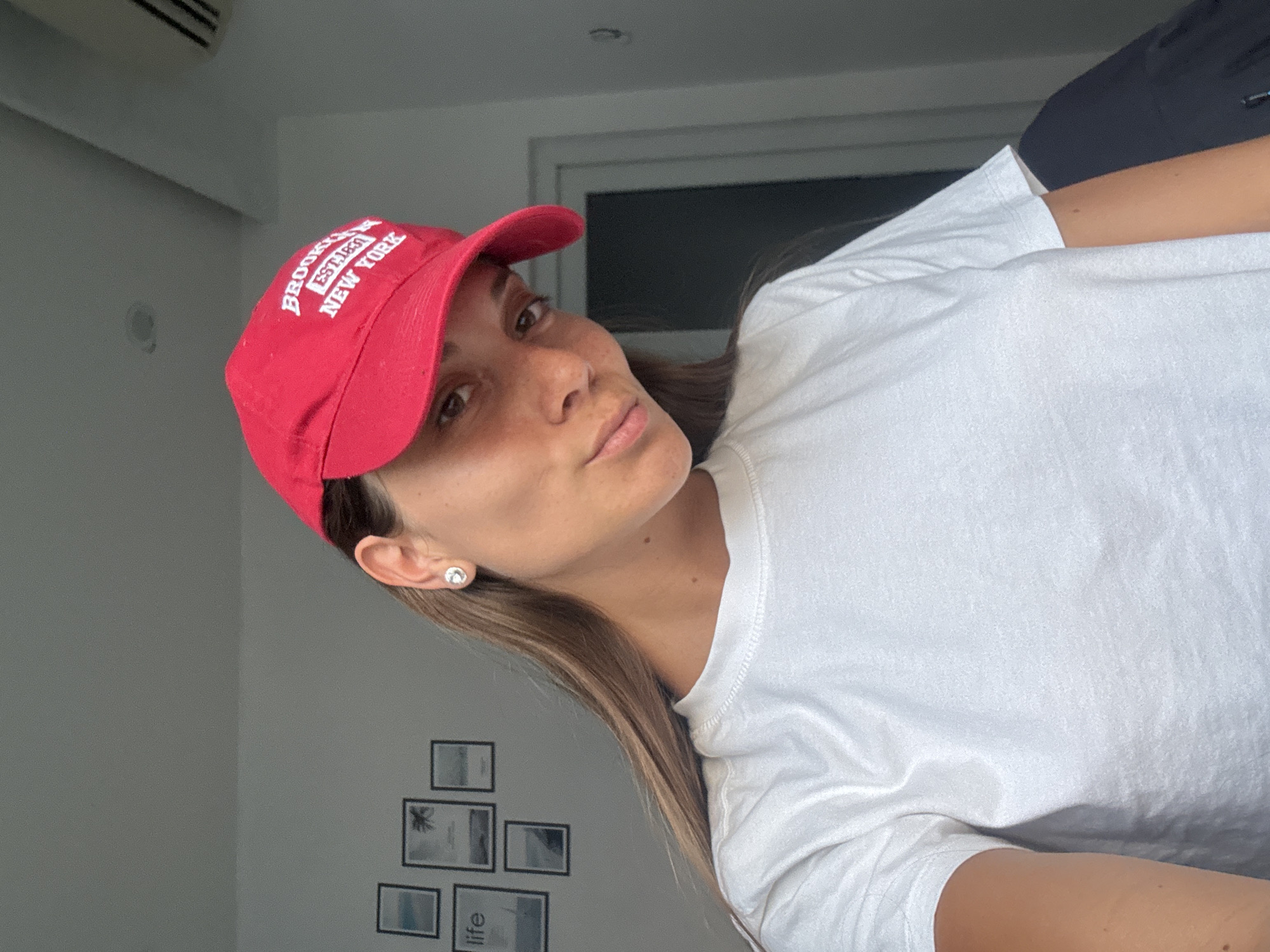 A woman wearing a red baseball cap that says "Brooklyn New York" and a white t-shirt is positioned in front of a white wall with framed pictures, possibly in an interior setting. Although the location is specified as Đà Nẵng, Vietnam, there is no visual evidence in the image to confirm this.