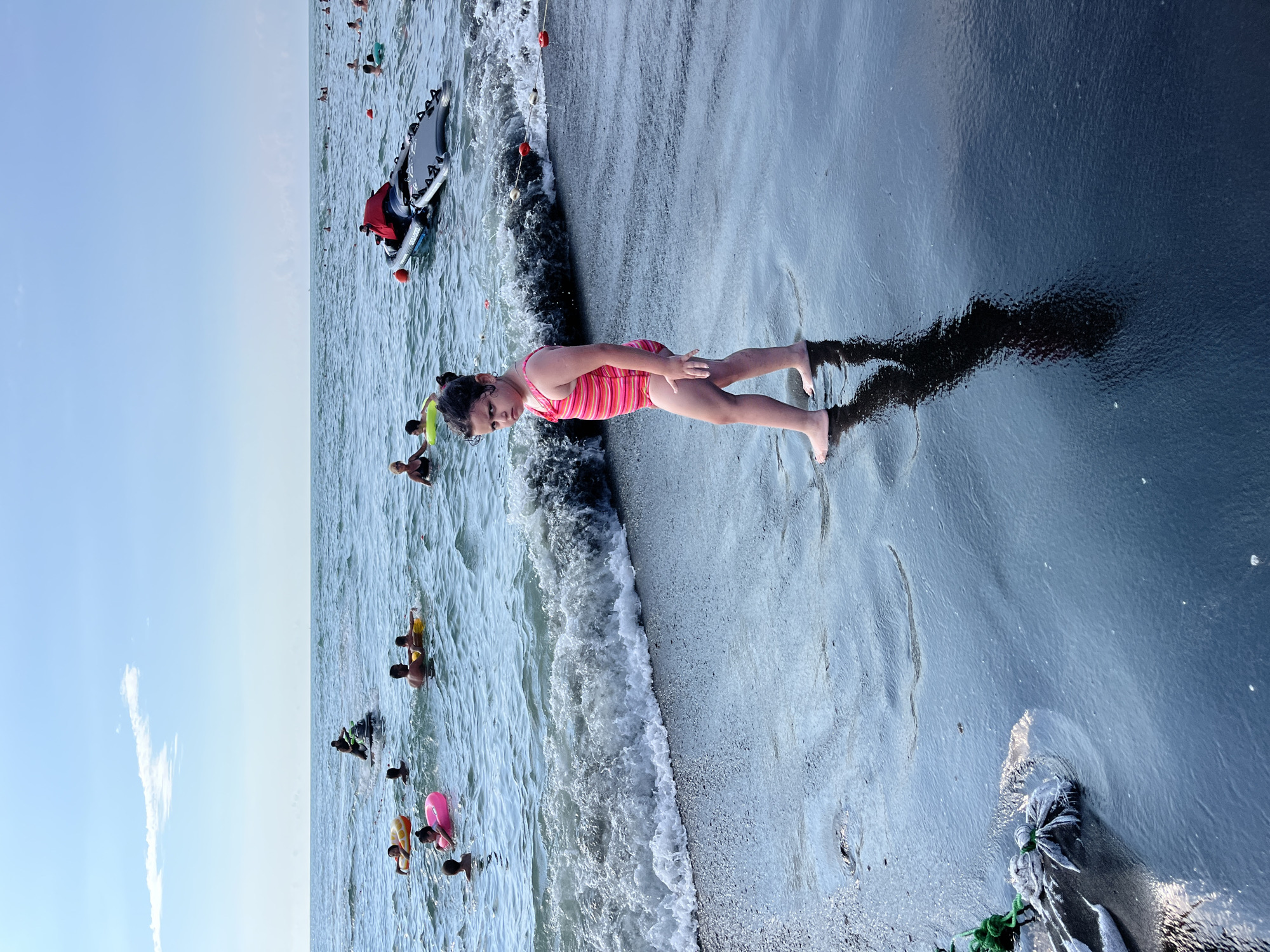The image depicts a beach scene in Ureki, Georgia, likely during the daytime given the bright sky. A young girl in a striped swimsuit stands on the wet sand near the water's edge, her shadow cast behind her. In the background, several people are visible in the water, some swimming, others on inflatable rings. A jet ski is also present in the water, with a person on it wearing a red vest. The water is choppy with small waves breaking on the shore. The sky is a clear blue with a few wispy clouds. The overall atmosphere suggests a recreational day at the beach.