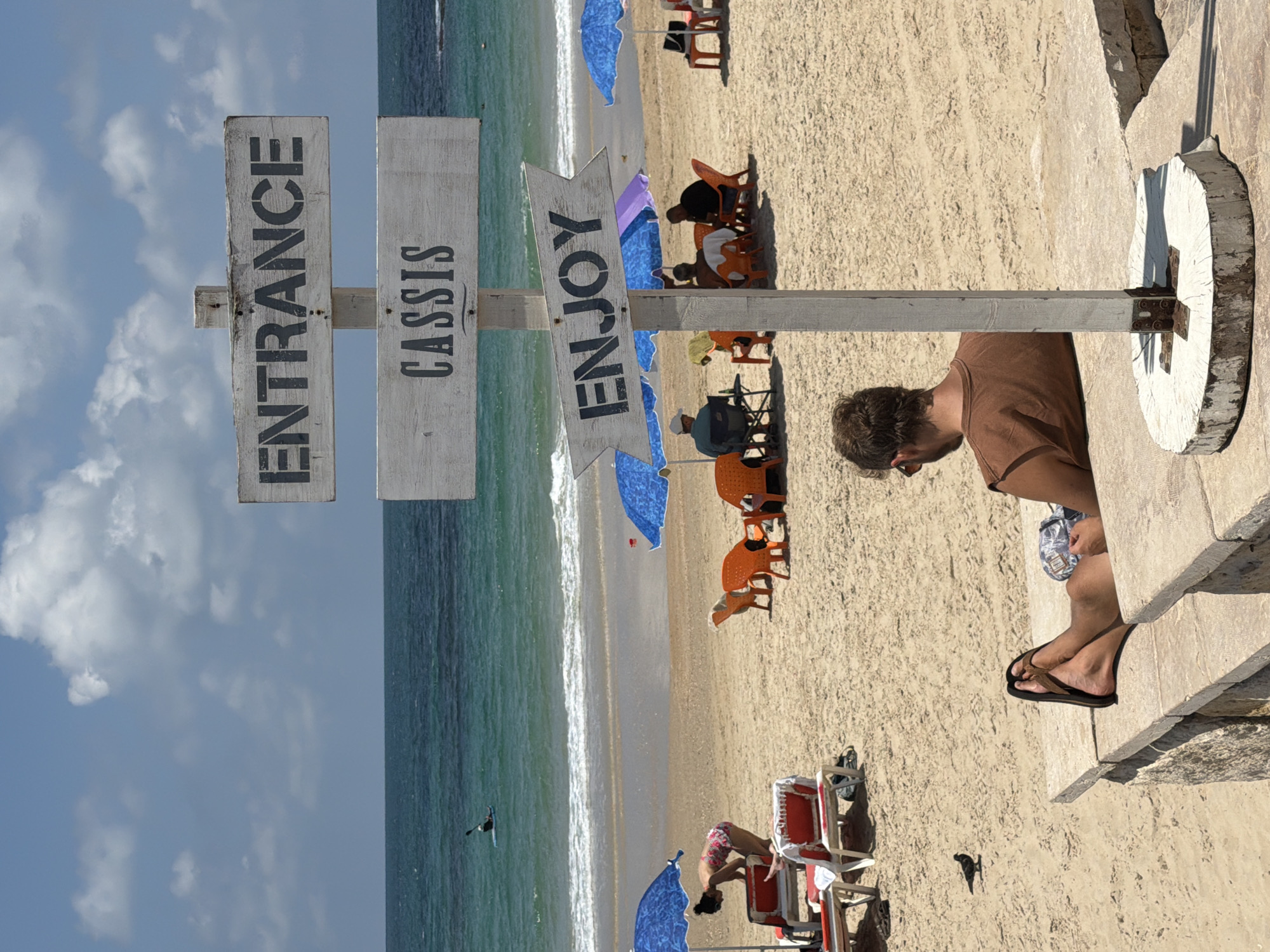 The image shows a beach scene in Cassis, France. A signpost with the words "Entrance," "Cassis," and "Enjoy" points toward the beach. People are relaxing in beach chairs and under umbrellas. A man is sitting on a concrete structure, facing toward the beach. The sky is partly cloudy.