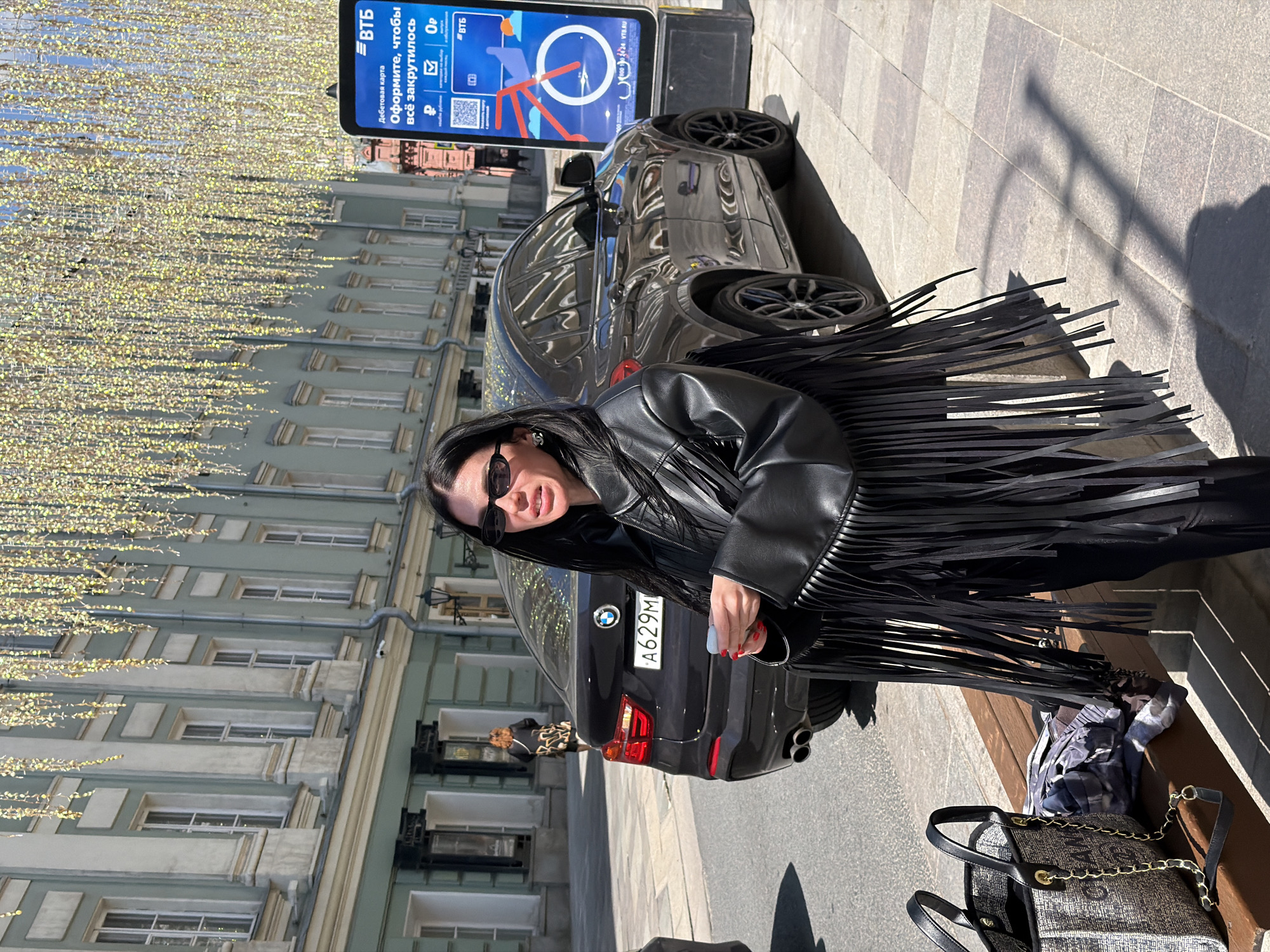 A medium shot of a woman with dark hair wearing a black, fringed leather jacket, black pants, and sunglasses, standing on a sidewalk next to a parked black BMW SUV with license plate "A629M." She is holding a white object in her hand. Behind the woman and the car is a light green and white building decorated with yellow string lights overhead. There is a VTB advertisement sign next to the car. A person can be seen walking in the distance on the sidewalk. A gray and white bag and a blue plaid shirt are sitting on a wooden bench beside the woman. The location is likely in Moscow, Russia, based on the presence of the VTB sign and the Russian license plate.