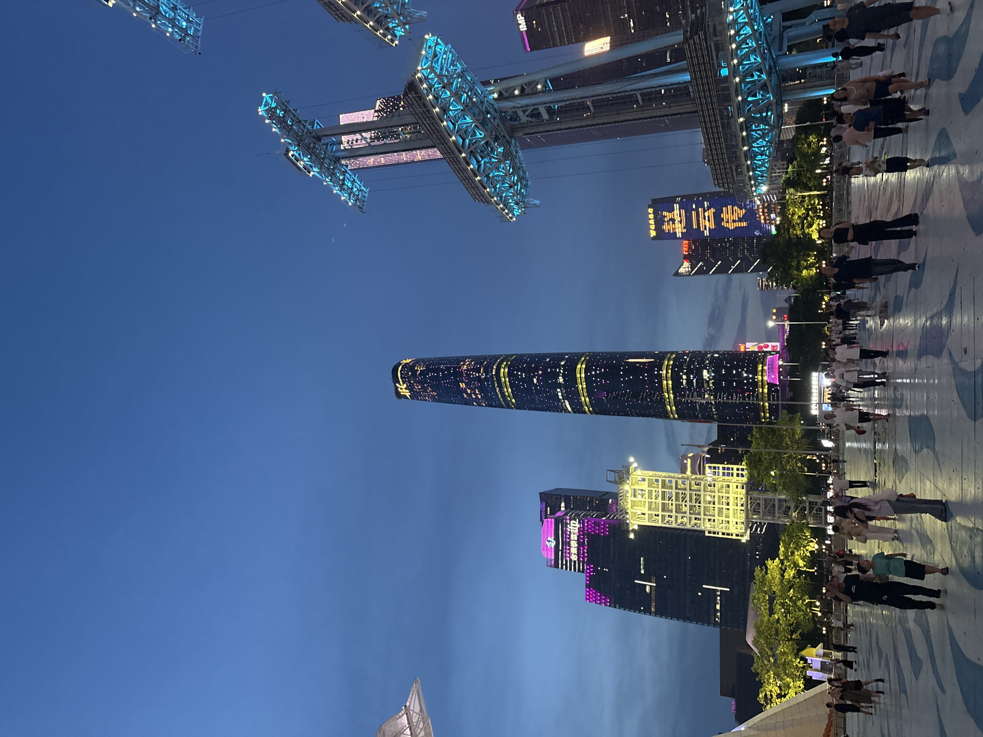 This is a nighttime shot of a bustling urban scene, likely taken in Tianhe District, China. The image is oriented vertically, with the sky forming the upper portion and a brightly lit pedestrian walkway in the lower portion.
The scene is dominated by several modern skyscrapers, illuminated with colorful lights, giving the impression of a vibrant cityscape at night. One prominent building is a tall, slender skyscraper with a pattern of lights that appears to be a spiral or a dynamic, flowing design. Another building features a large illuminated advertisement with Chinese characters, a combination of yellow and pink lights. There's also a structure that looks like a multi-story white trellis or scaffolding, also lit up. Overhead, what appears to be part of a large amusement ride or architectural installation is visible, adorned with blue lights.
The walkway below is paved with a patterned surface, possibly blue and white designs. A group of people are strolling along this walkway, engaged in casual activity. They are dressed in summer clothing, suggesting a warm evening. Their postures and arrangement indicate a relaxed, leisurely atmosphere. The time of day is clearly night, with the artificial lights of the buildings and the ride being the primary light sources, contrasting with the dark blue of the sky. The weather appears clear, with no visible clouds. The overall mood is one of urban energy and nighttime recreation.