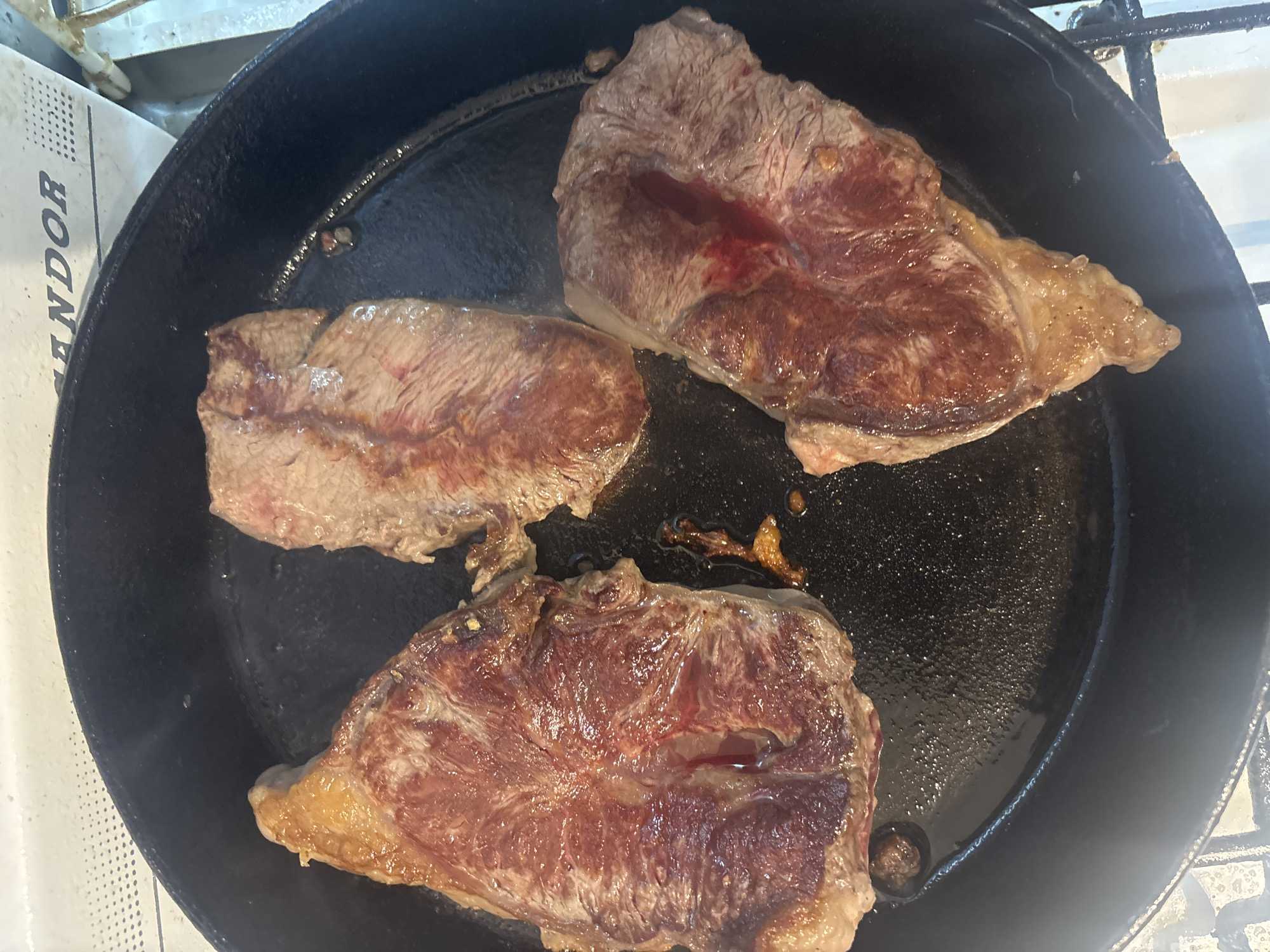 Three cuts of beef steak are cooking in a dark, circular cast iron pan, viewed from directly above. The steaks are raw to medium-rare on their visible surfaces, displaying reddish meat fibers, some marbling, and rendered fat around their edges. Liquid, likely a mixture of juices and cooking oil or rendered fat, is pooled in the pan around the steaks and coats their surfaces, giving them a glistening appearance. The pan itself is black and appears well-used, with some darker, crusty residue visible from previous cooking.
To the left of the pan, a portion of a white object with a textured or perforated surface is visible. Running vertically along this object are the capital letters "ANDOR", with the bottom part of the 'R' cut off. This object appears to be part of an appliance or counter. The overall setting suggests a kitchen environment. No people are visible, and the time of day and weather cannot be determined from the image. The scene captures an active cooking process.