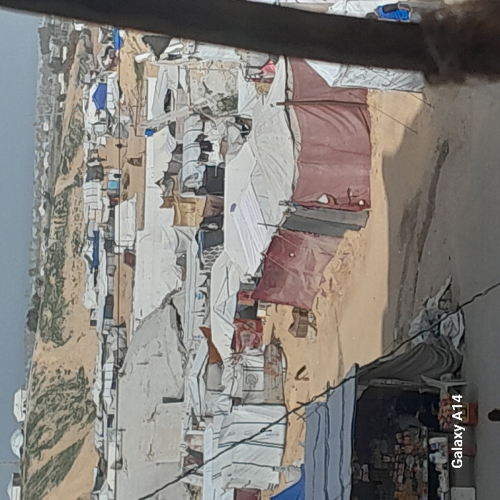 The image depicts a densely populated displacement camp in Khan Yunis, Palestinian Territory, under a hazy sky. The terrain is primarily sandy, with a prominent sandy hill covered in sparse vegetation visible in the background. Numerous makeshift shelters of various sizes, constructed from white tarpaulins, blue tarpaulins, and reddish-brown fabric, are tightly packed across the landscape. Laundry or fabrics are visible hanging to dry on some tents.
In the foreground, a small, dark-roofed kiosk or shop is open, displaying various goods on shelves. A person is seated inside this shop, and a white plastic chair is positioned nearby. Diagonal wires or cables traverse the immediate foreground. Scattered figures, mostly indistinct due to distance, are visible walking or standing among the tents and sandy pathways throughout the camp. A person wearing a blue garment is discernible near a white tent on the far right side of the frame. A dark, vertical object frames the right edge of the image. The environment suggests an improvised settlement with limited infrastructure.