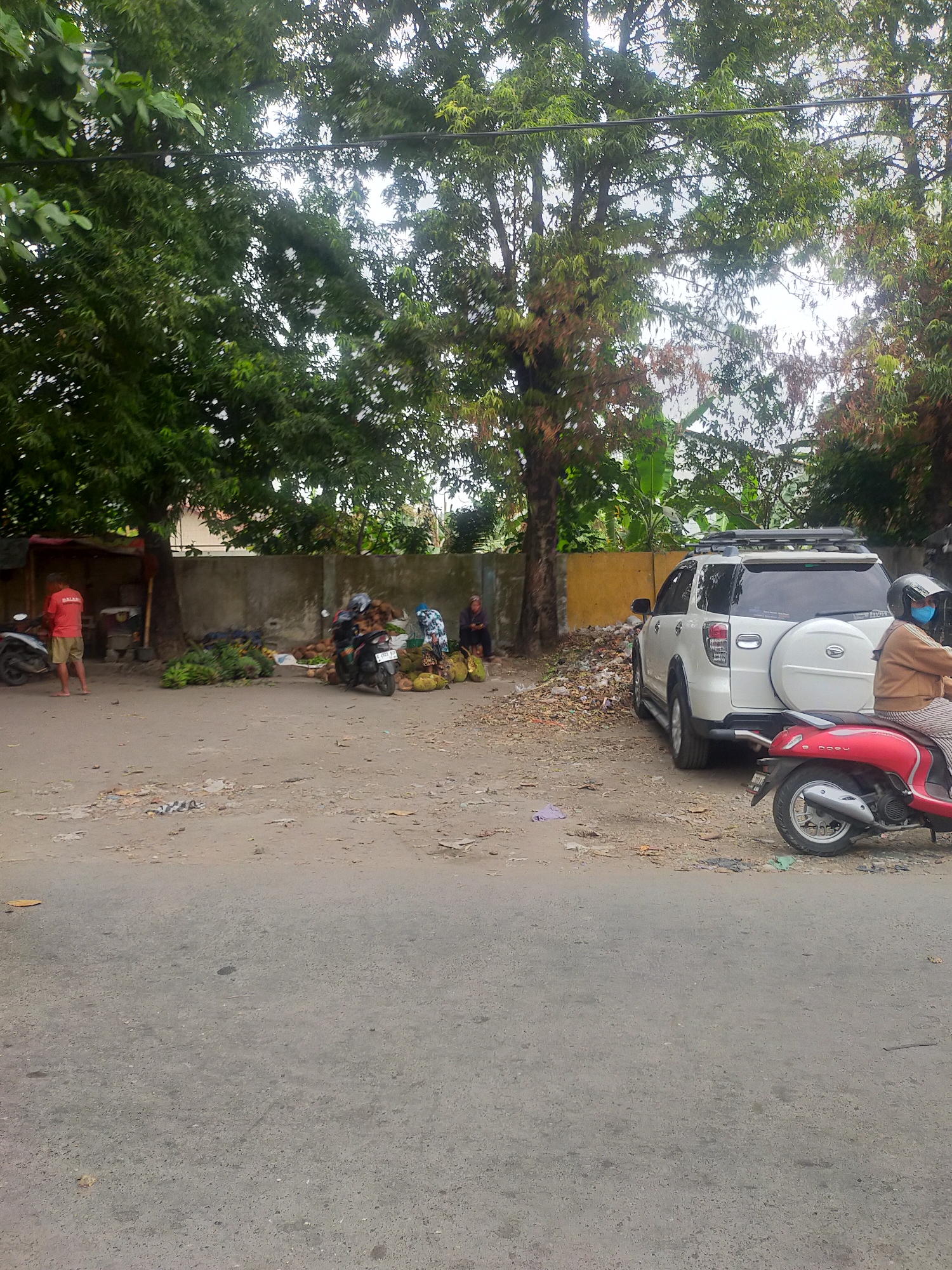 The image depicts a roadside market scene in Trayeman, Indonesia. A white SUV is parked on a dirt lot adjacent to a concrete wall. In the foreground, a red scooter with a rider wearing a blue mask and helmet is visible. Several people are seen at the market, some tending to piles of coconuts and bananas. A person in a red shirt stands near a motorbike on the left side of the image. The environment is characterized by a dusty, unpaved ground scattered with debris and foliage from surrounding trees.