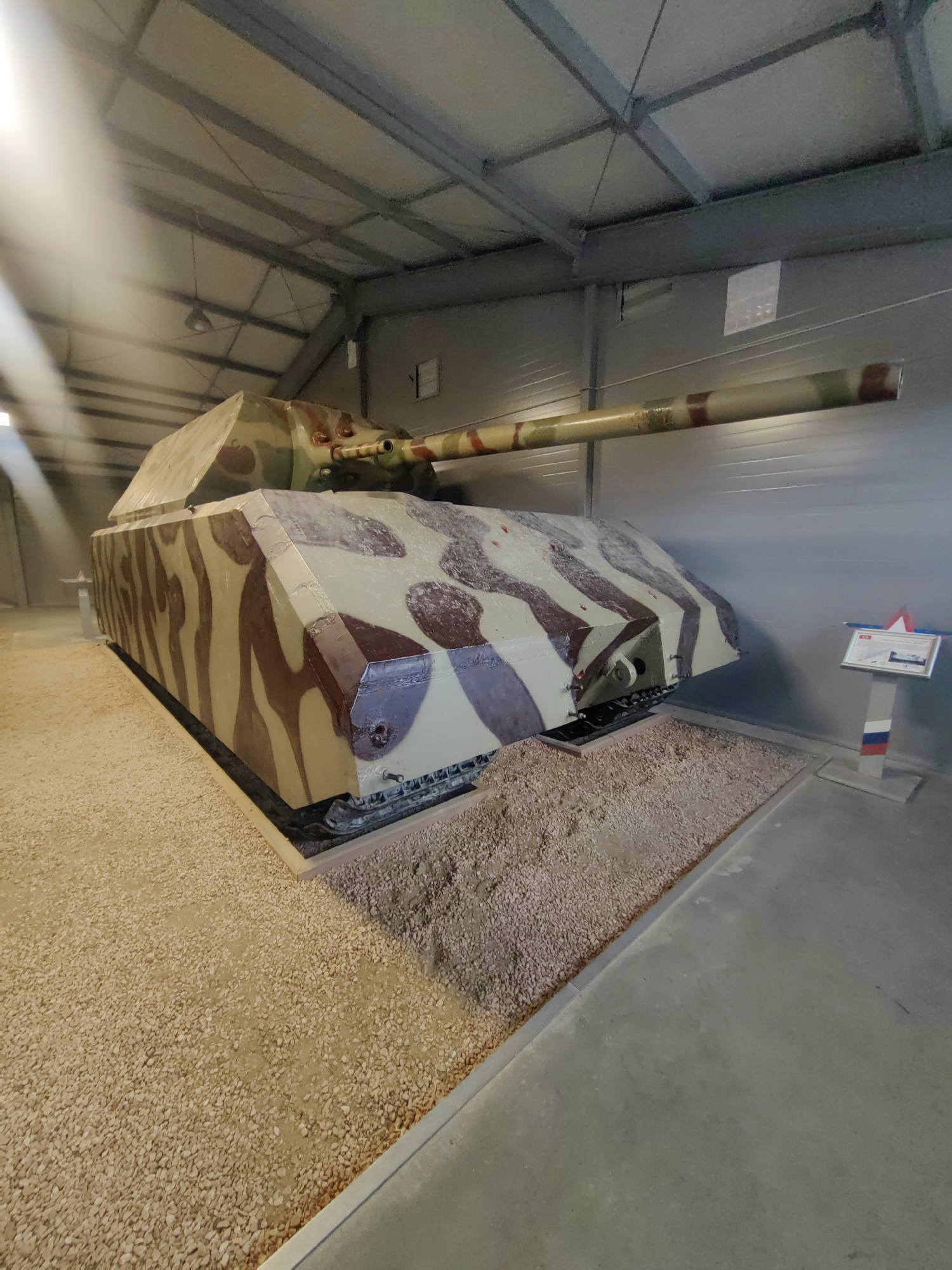 This media file depicts the Panzer VIII Maus, a German super-heavy tank from World War II, displayed indoors within a museum or exhibition hall. The tank is the sole subject of the image, positioned prominently in the center. It is painted in a camouflage scheme of brown and beige irregular shapes. The setting is a large, industrial-looking building with a high ceiling supported by metal beams and trusses. The floor around the tank is covered with gravel, creating a distinct display area.
A sign with a red and white star at the top and what appears to be the Russian flag colors on its base is visible to the right of the tank, suggesting a location in Russia, specifically Kubinka, known for its tank museum. The sign itself has some text and an image on a white background, though it is too small to be legible. Sunlight streams in from the left side of the building, creating bright highlights and casting shadows, indicating it is daytime. The overall atmosphere is that of a preserved historical artifact in a controlled exhibition environment. There are no people visible in the image. The focus is solely on the imposing presence of the tank.
