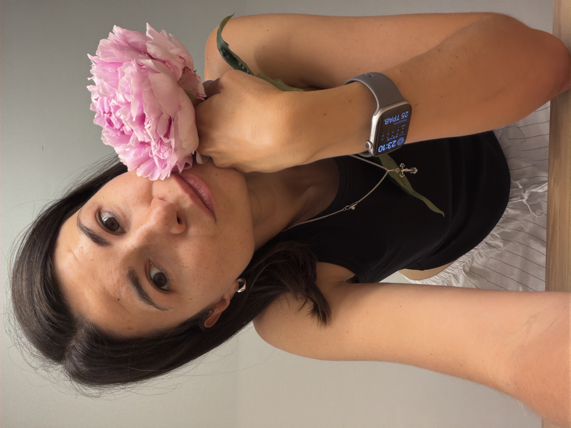 A woman is holding a large pink peony flower to her face. She is wearing a black tank top, a silver necklace with a cross pendant, and a grey smartwatch on her left wrist. The watch face displays the time as 23:10 and the date as 25 TBAB. The background is a plain light grey wall. Part of a vertically-striped garment, possibly shorts or a skirt, is visible near her arm. The image appears to be taken at an angle, possibly a selfie. There is no information to suggest that the location is Budva, Montenegro.