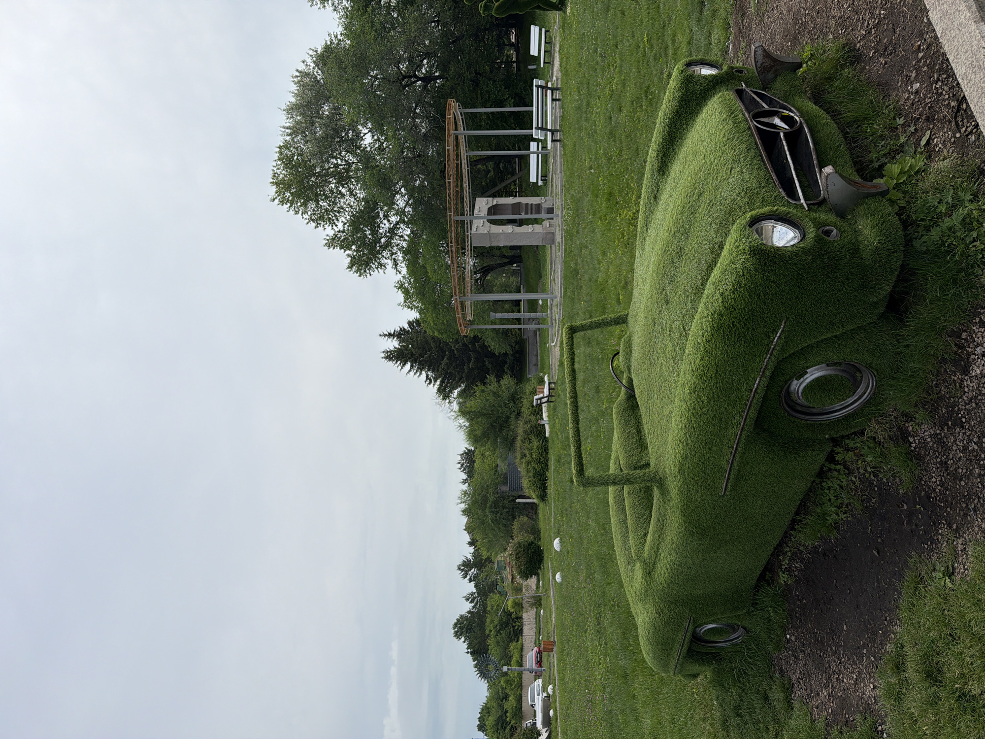 This is a photograph of a unique art installation in Birobidzhan, Russia, featuring a classic Mercedes-Benz convertible meticulously sculpted from living greenery. The car, covered in lush grass, is situated in a park-like setting with manicured lawns and mature trees. In the background, a circular pavilion with metal supports and a stone structure stands as a focal point. White benches are arranged symmetrically around the pavilion. Several white, spherical sculptures are dotted across the grass. The sky is overcast and a light gray. The overall scene depicts a public space designed for leisure and artistic appreciation, with the grass-covered car serving as the primary subject.