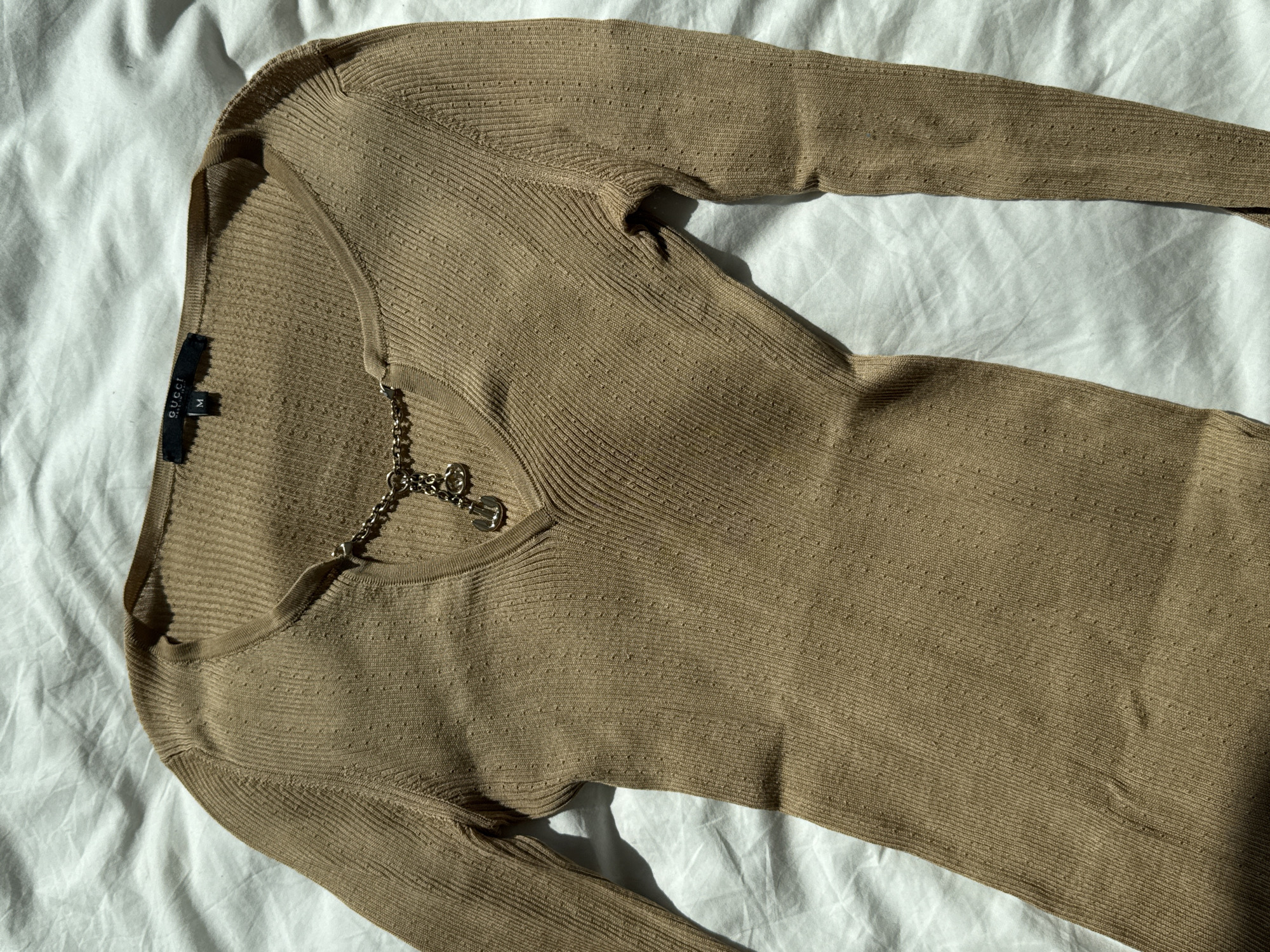 The image shows a light brown, long-sleeved, knit top laid out on a white sheet. The top features a silver chain embellishment at the neckline and a Gucci brand tag visible at the collar. The item is presented indoors under diffused natural light, suggesting a setting for product photography or display. The composition focuses solely on the garment, highlighting its texture and design details against the simple background. The setting is unknown, so a location cannot be verified from the image.