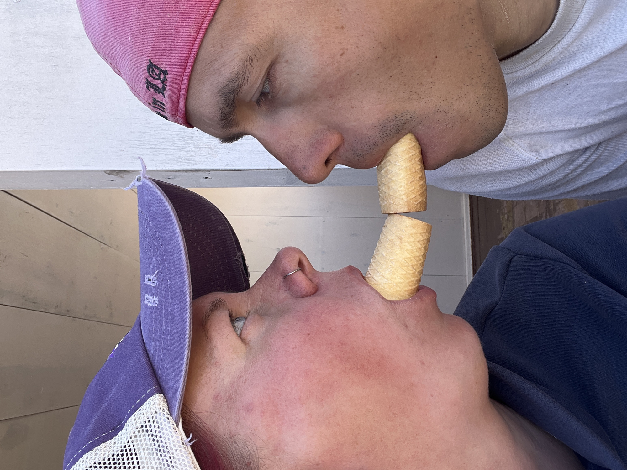Two people are positioned head to head, each with a waffle cone held between their lips, connected to form a single, longer cone. The person on the top wears a pink baseball cap and a white shirt. The person on the bottom wears a purple baseball cap and a blue shirt. The background appears to be a wooden structure, possibly a building. No specific details indicate the location is Chaikovsky, Russia.