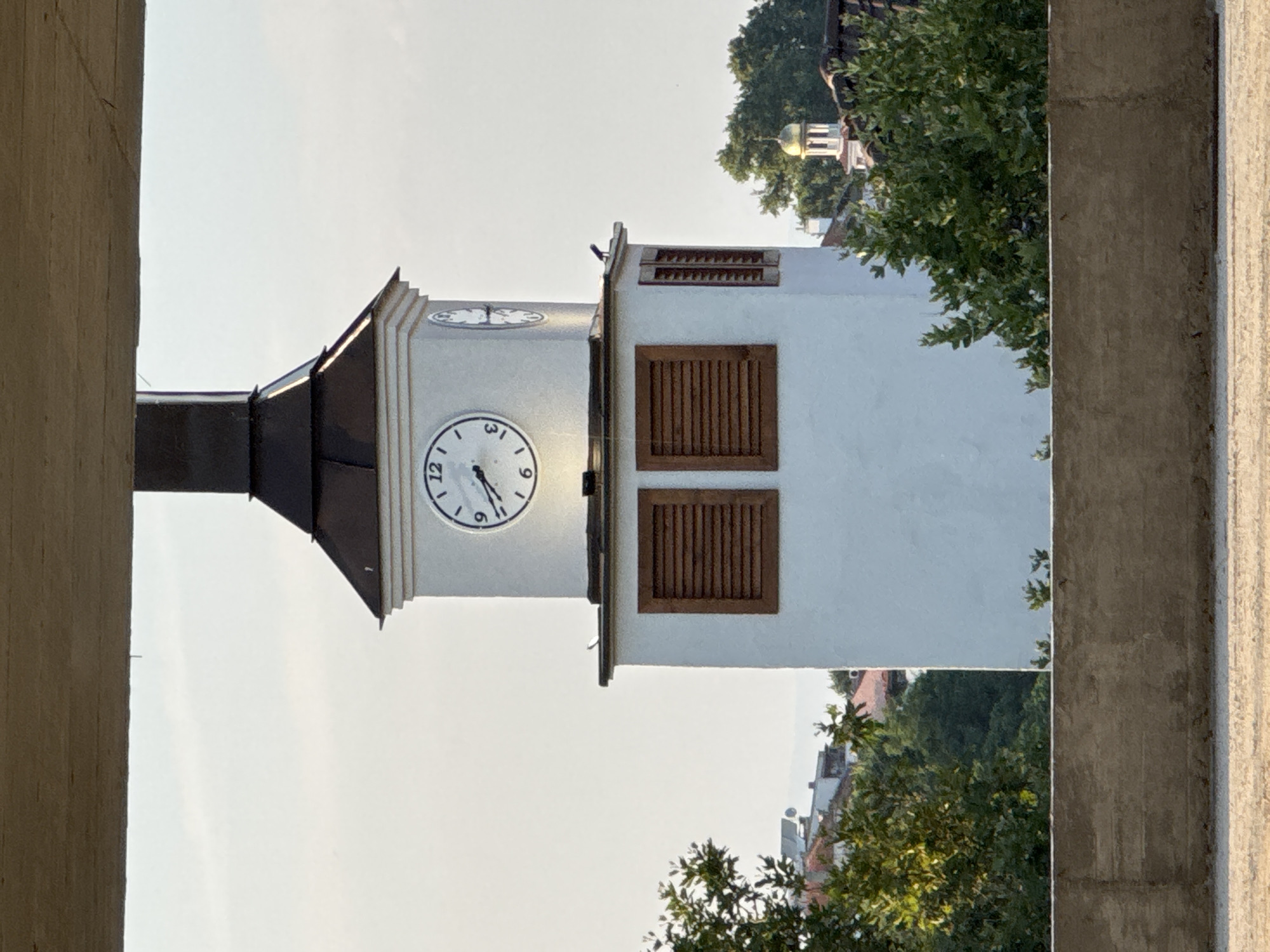 A white clock tower with a dark roof stands tall in Sozopol, Bulgaria. The tower has two visible clock faces, one displaying 9:35 and the other showing a time that is not clearly discernible. Below the clock faces are two windows with brown slatted shutters. In front of the tower, several green trees are visible, and in the background, the roofs of buildings can be seen, along with a small dome topped with a golden cupola. The scene is framed by a concrete structure at the bottom and a light-colored overhead structure. The sky is clear and pale blue.