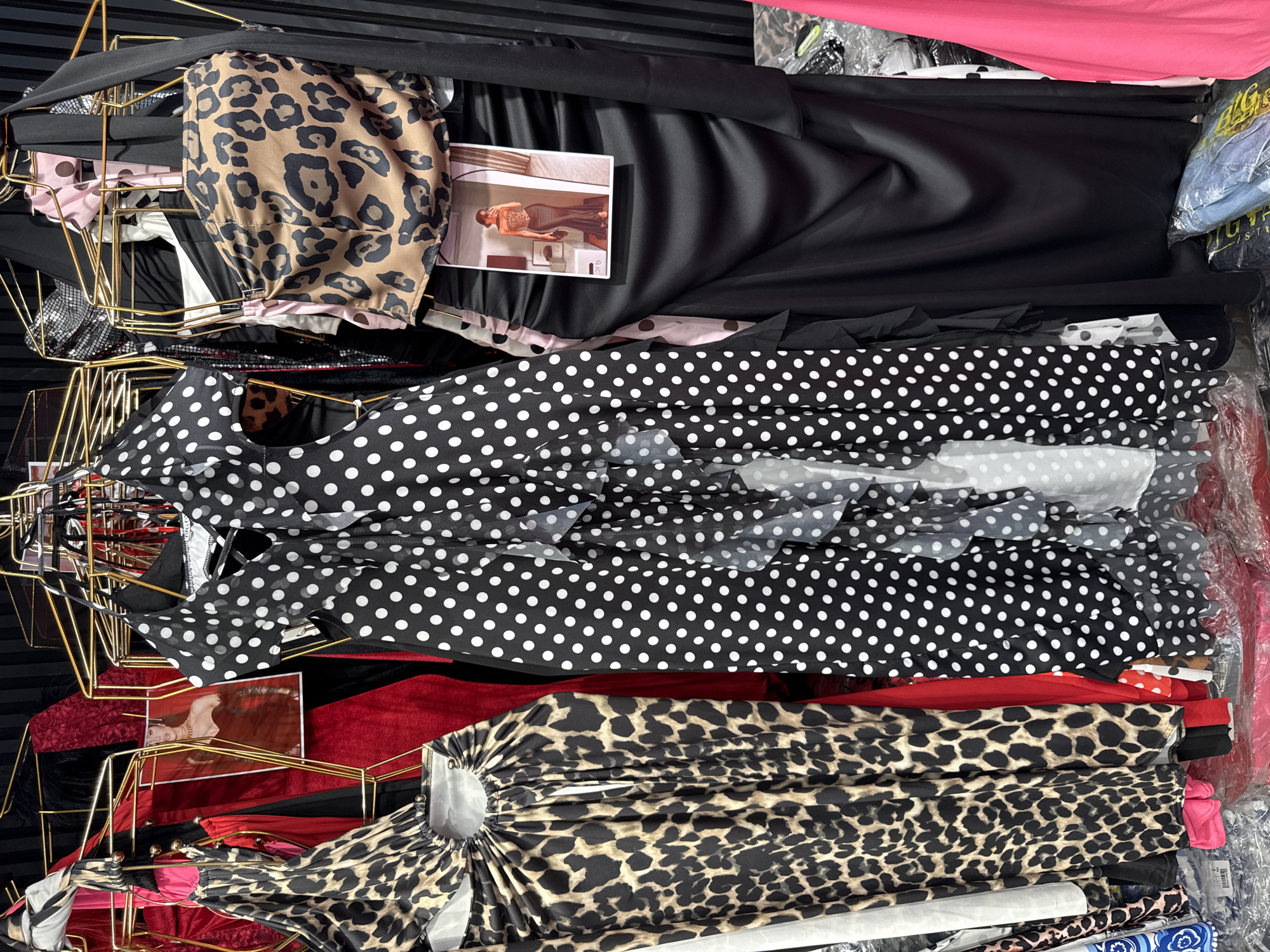 A display of women's clothing is shown, featuring several dresses and a leopard print top. The items are hung on gold-colored racks. In the center of the image, a black dress with white polka dots and ruffled detailing is prominent. To the left, a leopard print dress with a cutout detail is visible. To the right of the polka dot dress, a black satin skirt is displayed with a leopard print top hanging above it. A small promotional image of a woman wearing a similar outfit is attached to the black skirt. The background consists of a dark, possibly ribbed, surface. The clothing is presented in what appears to be a retail environment, possibly a store in the city of Gungoren, Turkey, given the context provided. No people are visible in the image.