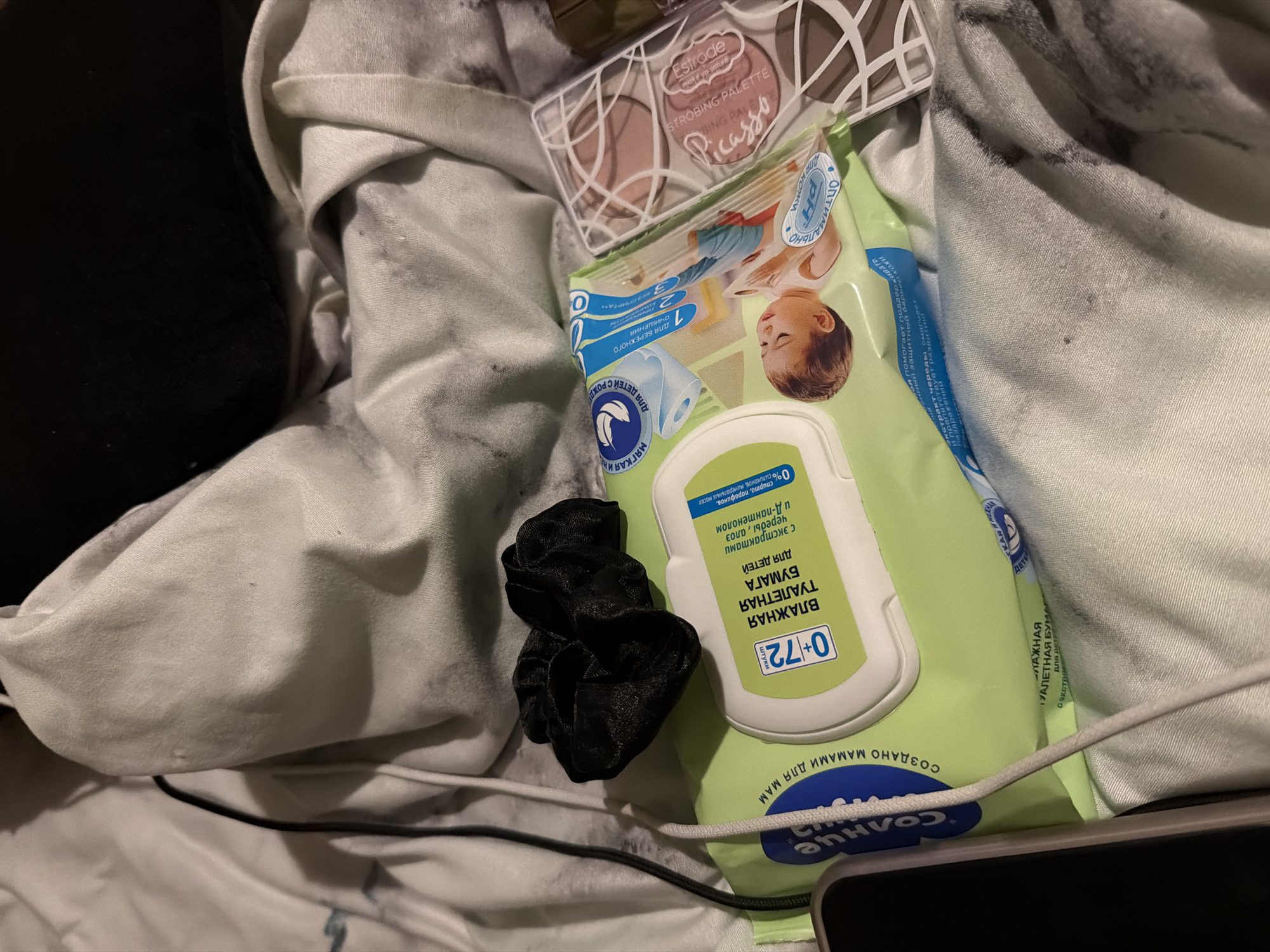 The image shows a close-up overhead shot of a pile of light-colored fabric, possibly a bed or couch, with a black scrunchie resting on a green package of wet wipes. To the right of the wet wipes is a makeup palette with circular pans of blush and highlighter. A phone is partially visible on the left side of the frame, and a white charging cable runs across the fabric. The lighting is dim, suggesting an indoor setting. There are no discernible city or location details.