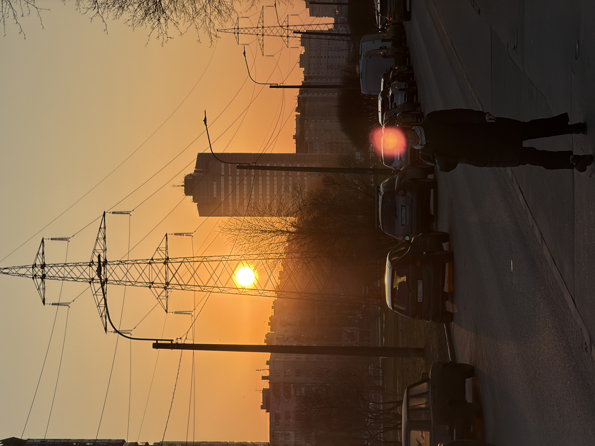 The image depicts a sunset scene in what appears to be a densely populated area. In the foreground, a person walks along a sidewalk lined with parked cars. The sky is dominated by a warm, orange glow from the setting sun. Power lines and utility poles cut across the horizon, adding a distinct urban element to the scene. Behind the power lines, several buildings and apartment complexes are visible, suggesting a residential or mixed-use area. Based on the general aesthetics, architectural style, and the presence of Russian license plates on the vehicles, it is inferred that the location is likely Saint Petersburg, Russia.