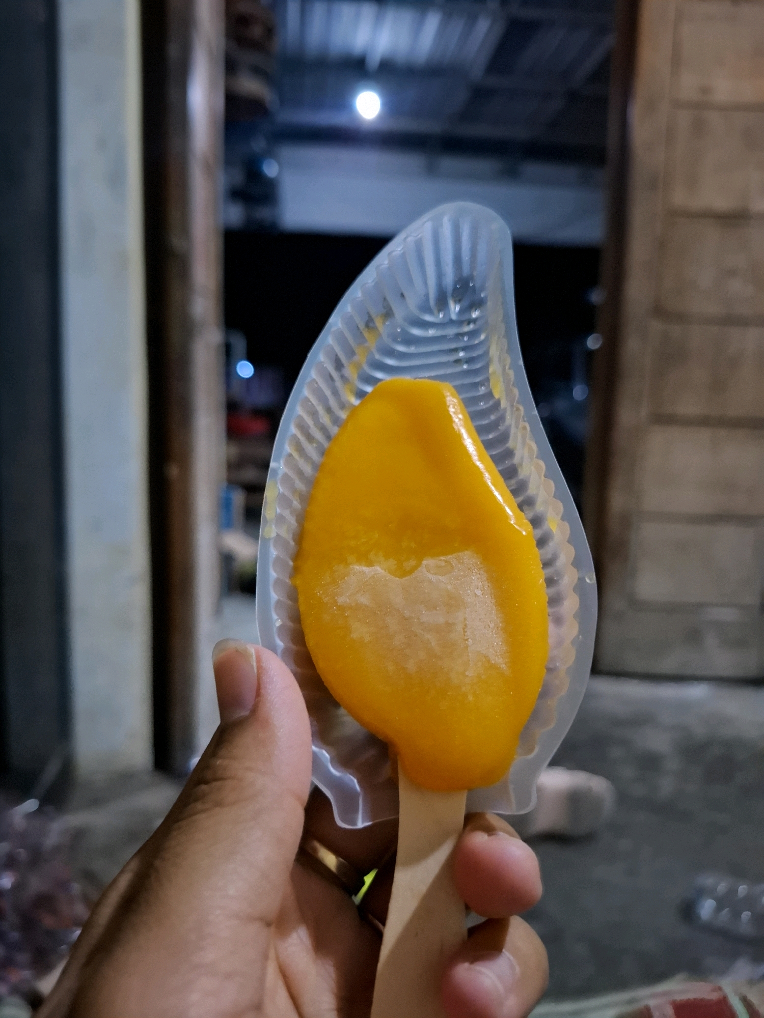 A person's hand holds a mango-flavored popsicle in a clear plastic mold. The popsicle is a vibrant yellow-orange and appears to be melting slightly. The background is blurred, showing a dimly lit outdoor setting with what appears to be wooden structures and possibly a roof overhead. The overall lighting suggests it is evening or night. The location is identified as Sembung, Indonesia.