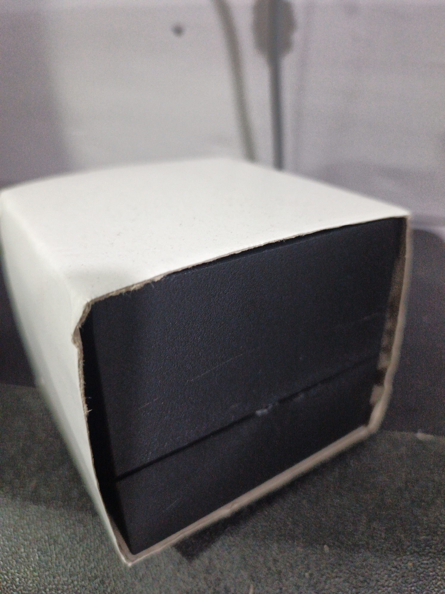A close-up, slightly angled shot shows a white cardboard box with a black inner box. The white outer box is partially opened and bent, revealing the edge of the black inner box. The black box appears to be made of a textured material. The background is out of focus and shows a light gray, textured wall or surface with shadows suggesting an indoor environment. The scene is devoid of people or any indication of activity. There is no information about the location in Kelapapati, Indonesia.
