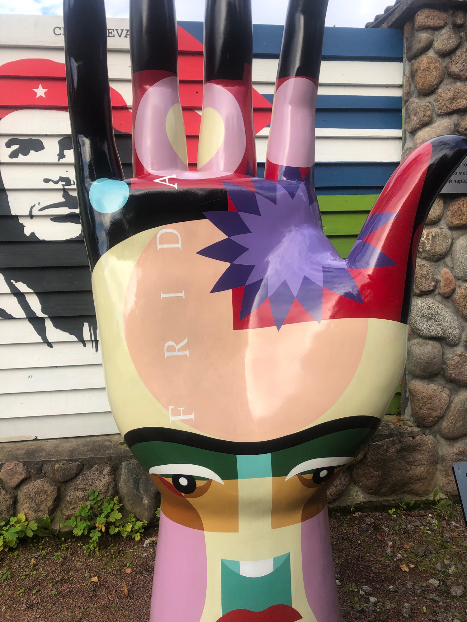 The image shows a colorful, larger-than-life sculpture of a hand, painted with abstract facial features on its palm and fingers. The sculpture is positioned outdoors, in front of a wall composed of horizontal wooden planks in alternating white and blue stripes, and a section of rough, natural stone wall on the right. To the left of the sculpture, there is a mural of a face with black and white stripes. The ground is covered in dark brown gravel with scattered dry leaves and small green plants. The sculpture itself features a yellow-toned palm area with the word "FRIDA" vertically inscribed in white letters. The fingers are black with various colored abstract shapes and facial elements painted on them, including eyes and lips. The sculpture's base has a stylized face with green eyebrows, brown and white eyes, and pink and red lips. The overall scene suggests an outdoor art installation or public art display, possibly in a park or recreational area.