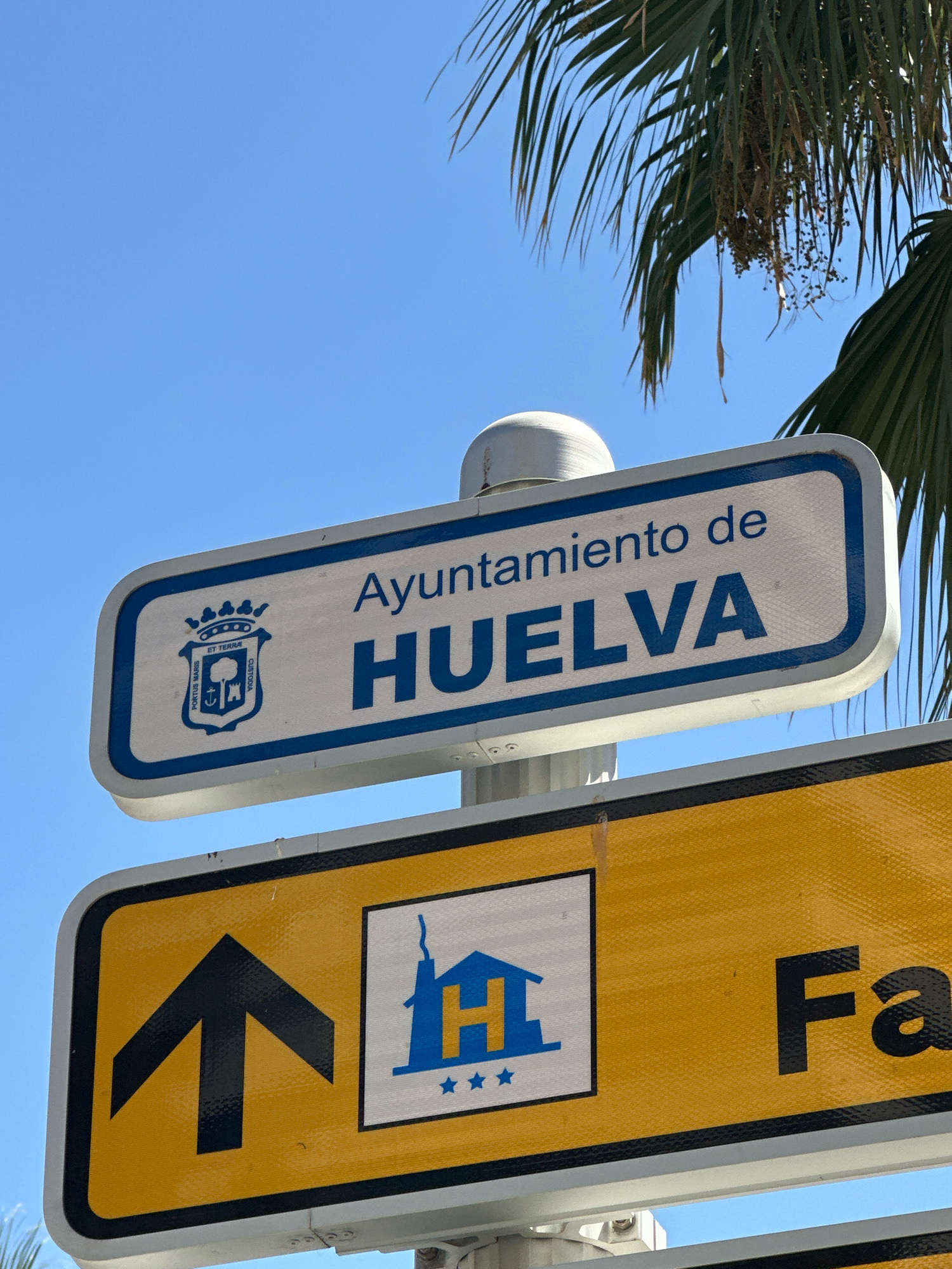 A sign for the Ayuntamiento de Huelva (City Hall of Huelva) is visible against a clear blue sky. The sign features the city's coat of arms on the left and "Ayuntamiento de HUELVA" written in blue letters on a white background with a blue border. Below this, a yellow directional sign with a black border displays a black upward arrow on the left, a white square with a blue house symbol and three blue stars in the center, and the partial text "Fa" in black on the right. Palm fronds are visible at the top right of the image.