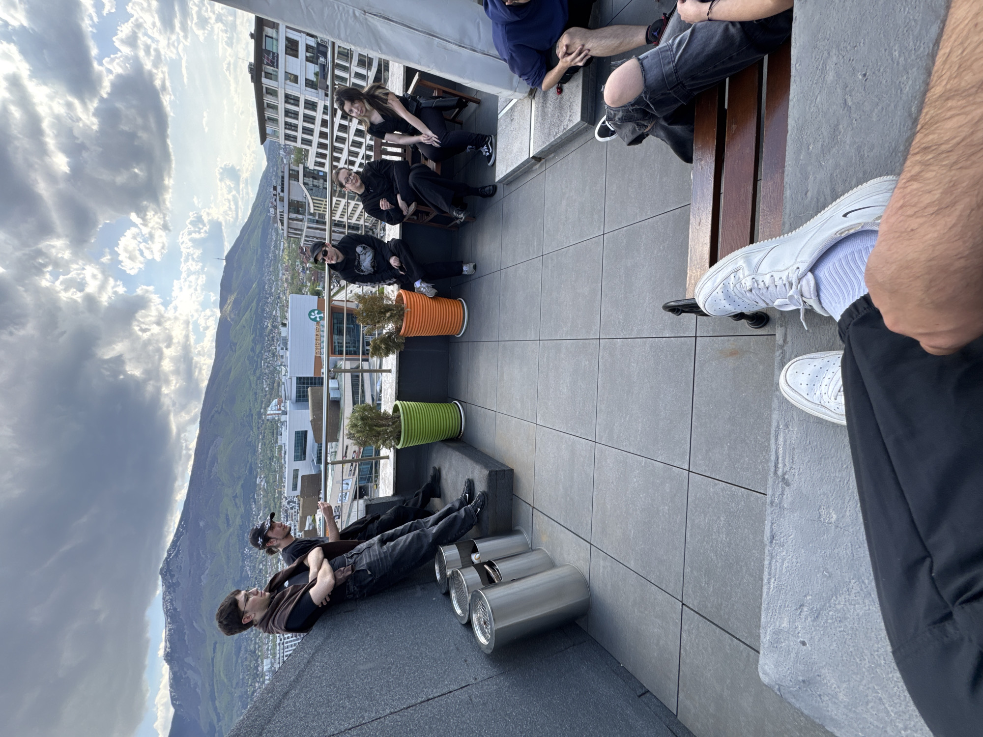 An outdoor scene captured from a high vantage point, likely a rooftop or large balcony in Sofia, Bulgaria, shows a group of people relaxing under a bright, cloudy sky. The image is oriented vertically (or 90 degrees rotated counter-clockwise).
In the foreground, to the left, two young men are lounging casually on a dark grey ledge or raised platform, facing towards the mountainous landscape in the distance. One wears a brown top and glasses, while the other has a black cap. Nearby, several cylindrical metallic objects are arranged on the grey tiled floor.
Towards the mid-ground and right, a larger group of individuals is seated and lounging. One person in the immediate foreground, partially cropped, wears white sneakers and dark pants. Further back, two individuals are seated on a wooden bench, one with ripped black jeans. Adjacent to them, three people are seated on chairs around a table; a woman with long dark hair, dressed in black, smiles towards the camera, while two others in dark clothing are seated nearby. Two large potted plants, one orange and one green, are positioned near this group, adding color to the tiled floor.
The setting overlooks an urban area with buildings, giving way to a prominent mountain range in the background. The sky is bright with soft, scattered clouds. A white sign with a green clover-like symbol and partially obscured text (possibly "BIZNES TSENTUR") is visible on a building or railing in the mid-ground. The overall mood is relaxed and social, with individuals enjoying the view and company. The time of day appears to be late afternoon or early evening due to the lighting.