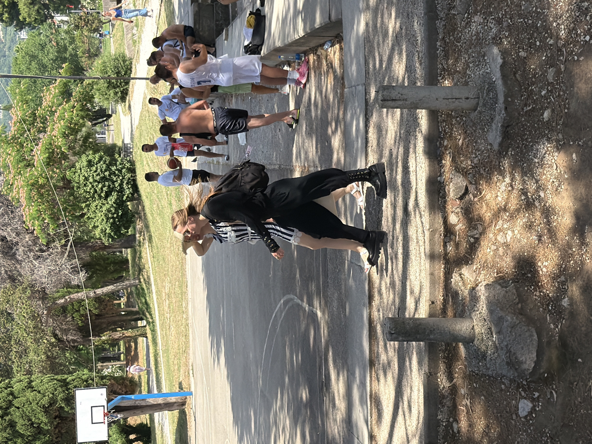 In Budva, Montenegro, a group of individuals are gathered near a basketball court on a sunny day. In the foreground, two women are walking across a paved area, one wearing a black and white striped dress and the other in black trousers and a black top. The woman in the striped dress has blonde hair and is wearing sandals, while the woman in black is wearing black boots and carrying a backpack. Several men are visible in the background, some of whom are playing basketball or watching. One man is holding a basketball, and others are wearing athletic clothing. A basketball hoop and backboard are set up on the court. The surrounding environment includes trees, grass, and a paved pathway. The scene appears to be an outdoor recreational area.