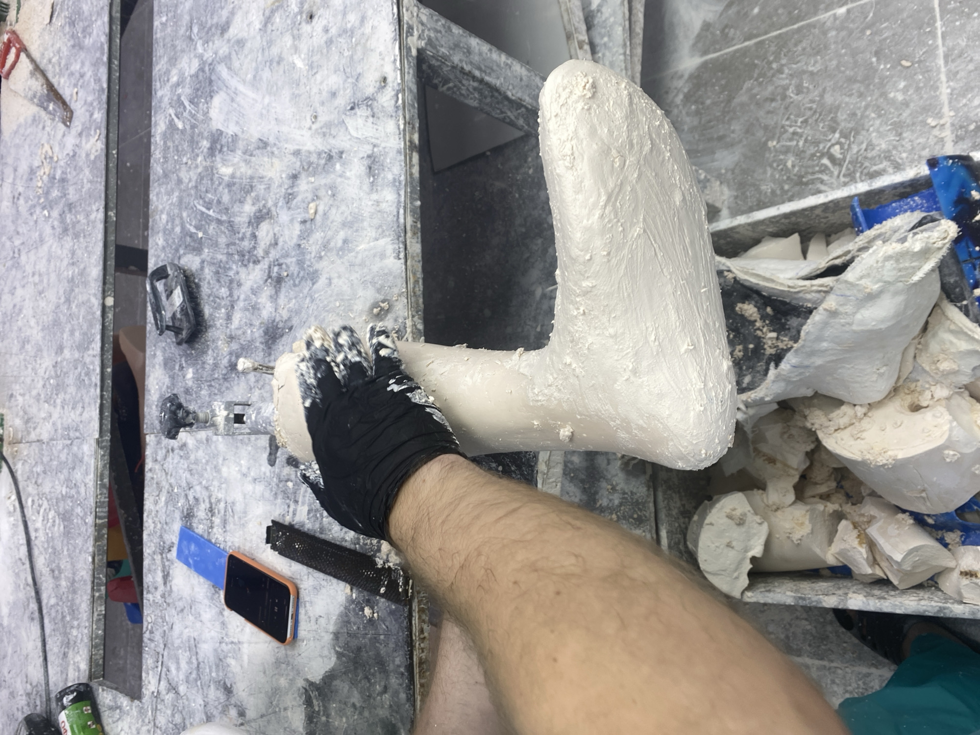 The image shows a close-up, overhead view of a person's leg and foot being cast with plaster. The subject is wearing a black glove on their right hand, which is applying the plaster to the foot. The foot is already partially covered in a thick, white plaster. The scene appears to be a workshop or studio, with various plastering tools and materials visible in the background. There are bins filled with broken plaster casts and a blue plastic spatula. The surfaces are dusty and appear to be made of concrete or similar material. The lighting is bright, suggesting an indoor environment. There is no clear indication of the time of day or weather conditions. The overall impression is one of a practical, hands-on activity. No text is visible in the image.