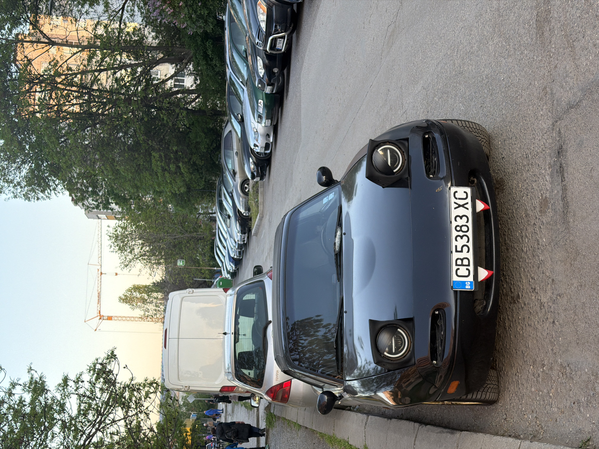 The image shows a street scene in Sofia, Bulgaria. A black car with the license plate "CB 5383 XC" is parked on the right. A white van and several other cars are parked behind it. There are trees on either side of the street. In the background, a building can be seen behind the trees, and there is a construction crane visible further back. A group of people are walking on the sidewalk near the white van.