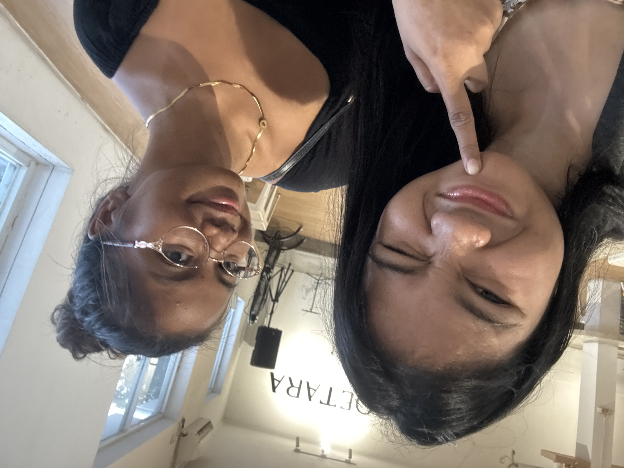 Two women are in the frame. The woman on the left wears round glasses and has her hair in a bun. She is also wearing a gold necklace. The woman on the right has dark hair and is pointing at her cheek. The background shows a room interior, possibly a shop, with the word "SETARA" visible on the ceiling. The location is North Jakarta, Indonesia.