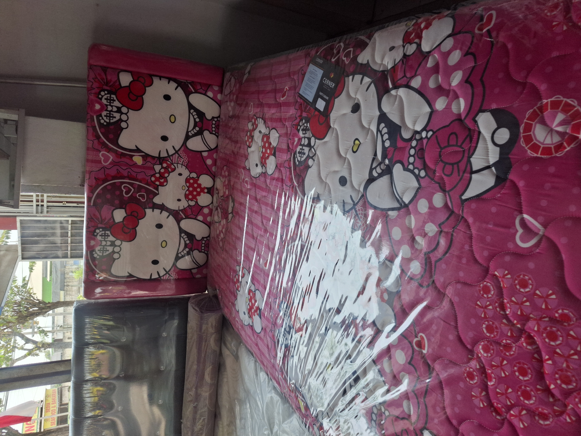 A brightly lit indoor store in Banjarmasin, Indonesia, displays a children's mattress with a Hello Kitty theme. The mattress features a pink comforter with multiple images of Hello Kitty characters in various poses and outfits. The headboard, also pink, is adorned with larger images of Hello Kitty. The mattress is covered in a transparent plastic wrap, reflecting the surrounding light. Next to the Hello Kitty mattress is a dark-colored mattress with a tufted headboard, also wrapped in plastic. In the background, a sign with Indonesian text and a red and white flag are visible through a window, suggesting the store is open for business. The environment appears to be a retail space selling bedding or furniture.