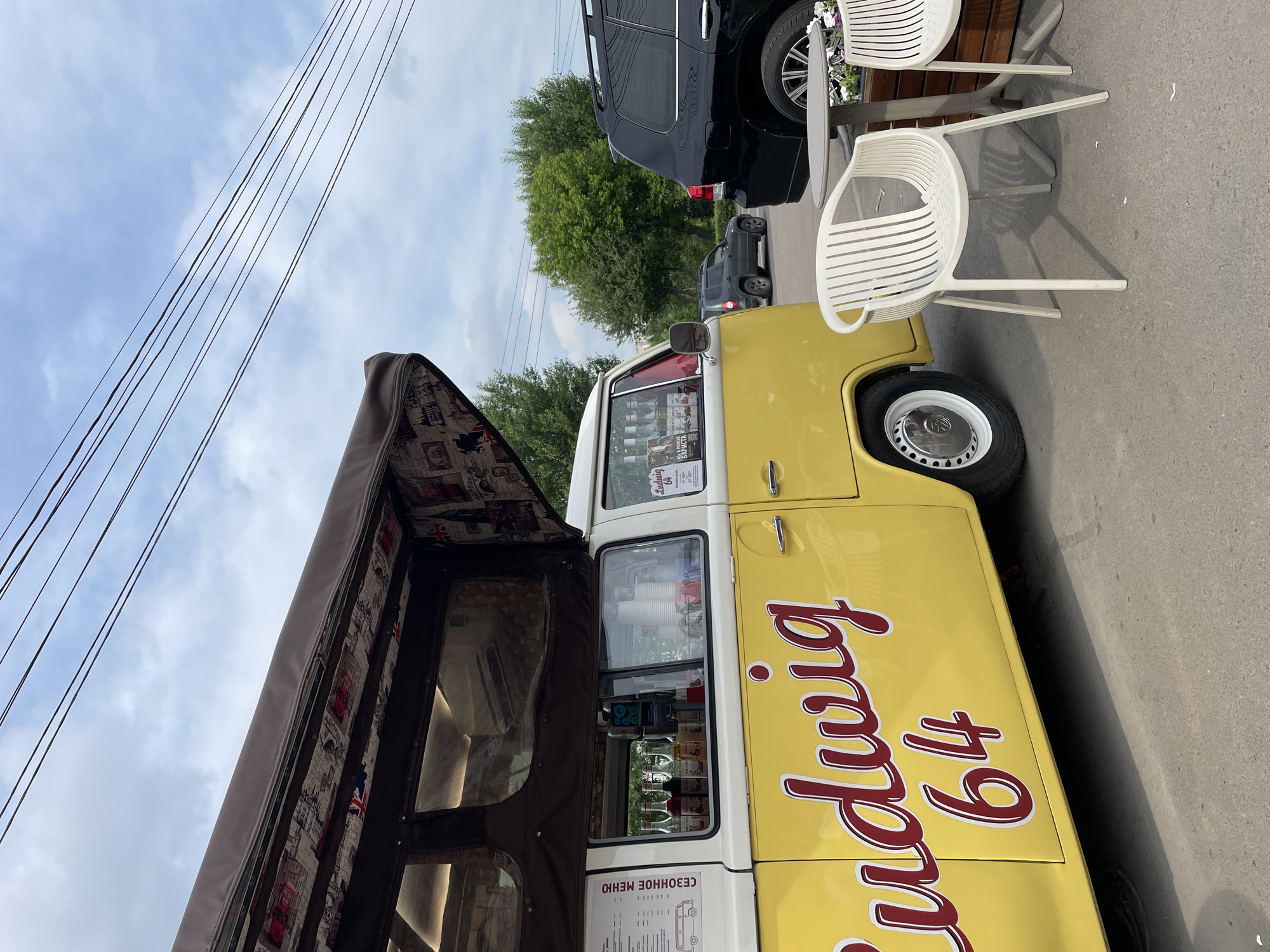 A yellow Volkswagen Type 2 van, identified as "Ludwig 64" on its side, is parked on an asphalt surface. The van has an open awning decorated with a London-themed pattern. Through the windows, bottles of syrup and stacks of paper cups are visible. To the right of the van, a small round table and two white plastic chairs are set up. Behind them is a wooden planter box with flowers and a dark SUV. In the background, a smaller grey SUV is driving on a road lined with green trees. Overhead, power lines crisscross against a cloudy sky. The image appears to be taken from a slightly low angle, looking towards the van and the seating area. This scene is located in the city of Krasnoyarsk, Russia, likely in an outdoor commercial area or park.