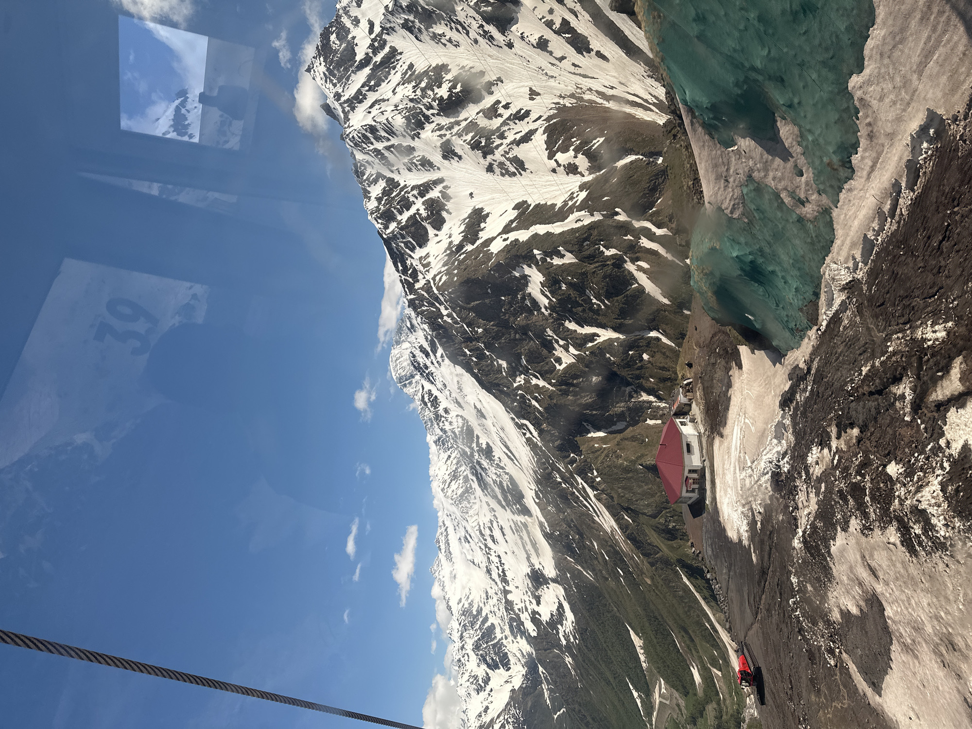 The image captures a scenic view of the snow-capped Caucasus Mountains, likely from a cable car, with the town of Terskol, Russia visible in the background. In the foreground, a red snow groomer is parked on a dark, rocky slope. A small white building with a red roof is situated on the terrain, and in the distance, several figures are visible near the building. The mountains are covered in snow with patches of dark rock and green vegetation at lower elevations. The sky is blue with scattered white clouds. Reflections on the glass surface show a number "39" and a silhouette of a person.