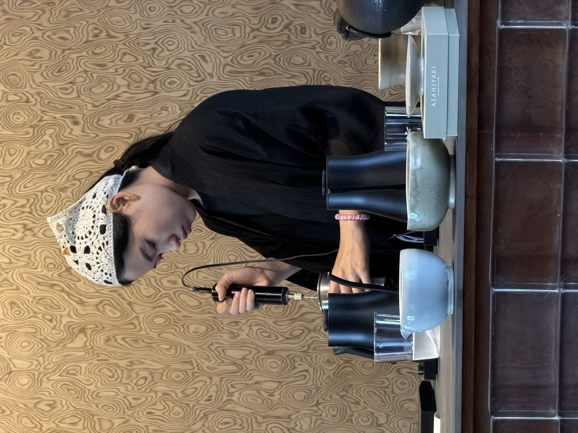 A young Asian woman wearing a black kimono-style robe and a white crocheted head covering is standing behind a dark brown counter. Her dark hair is pulled back in a ponytail. She is holding a black electric whisk-like device with a silver tip over a silver bowl, appearing to be whisking something. To her left is a clear glass with a clear liquid and a black pitcher. In front of her, from left to right, are a light blue ceramic bowl, a pale green ceramic bowl, and a light blue box with "ASAHIYAKI" written in white lettering. The background is a wooden wall with a swirling, wood-grain pattern. The counter itself has a tiled, reflective surface. The scene appears to be set in an indoor establishment, possibly a cafe or shop, in Chiang Mai City Municipality, Thailand, indicated by the product packaging.