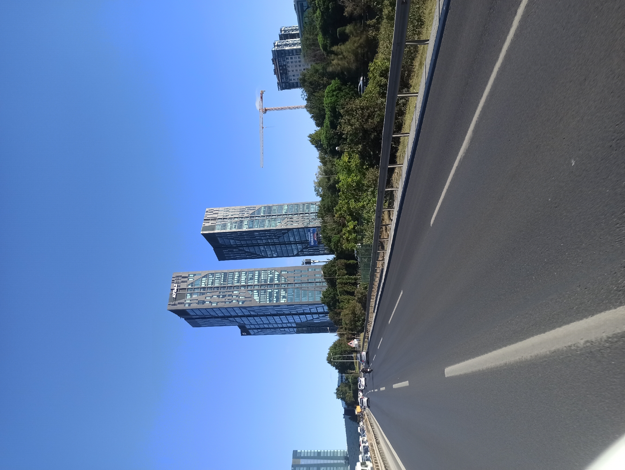 The image depicts a view from a highway in Ümraniye, Turkey, looking towards a cluster of modern, tall buildings. Two prominent skyscrapers, likely office buildings, dominate the skyline. The building on the left has "AYGAZ" inscribed at its top. To the right of these, a construction crane is visible, indicating ongoing development. In the foreground, the asphalt road with white lane markings stretches into the distance, with several vehicles traveling on it. A metal guardrail lines the side of the highway, separating it from a dense area of green trees and foliage. The sky is a clear, bright blue, suggesting a sunny day. Another building, appearing to be a residential complex, is visible in the far right of the frame. The overall scene suggests an urban environment characterized by both established modern architecture and active construction.