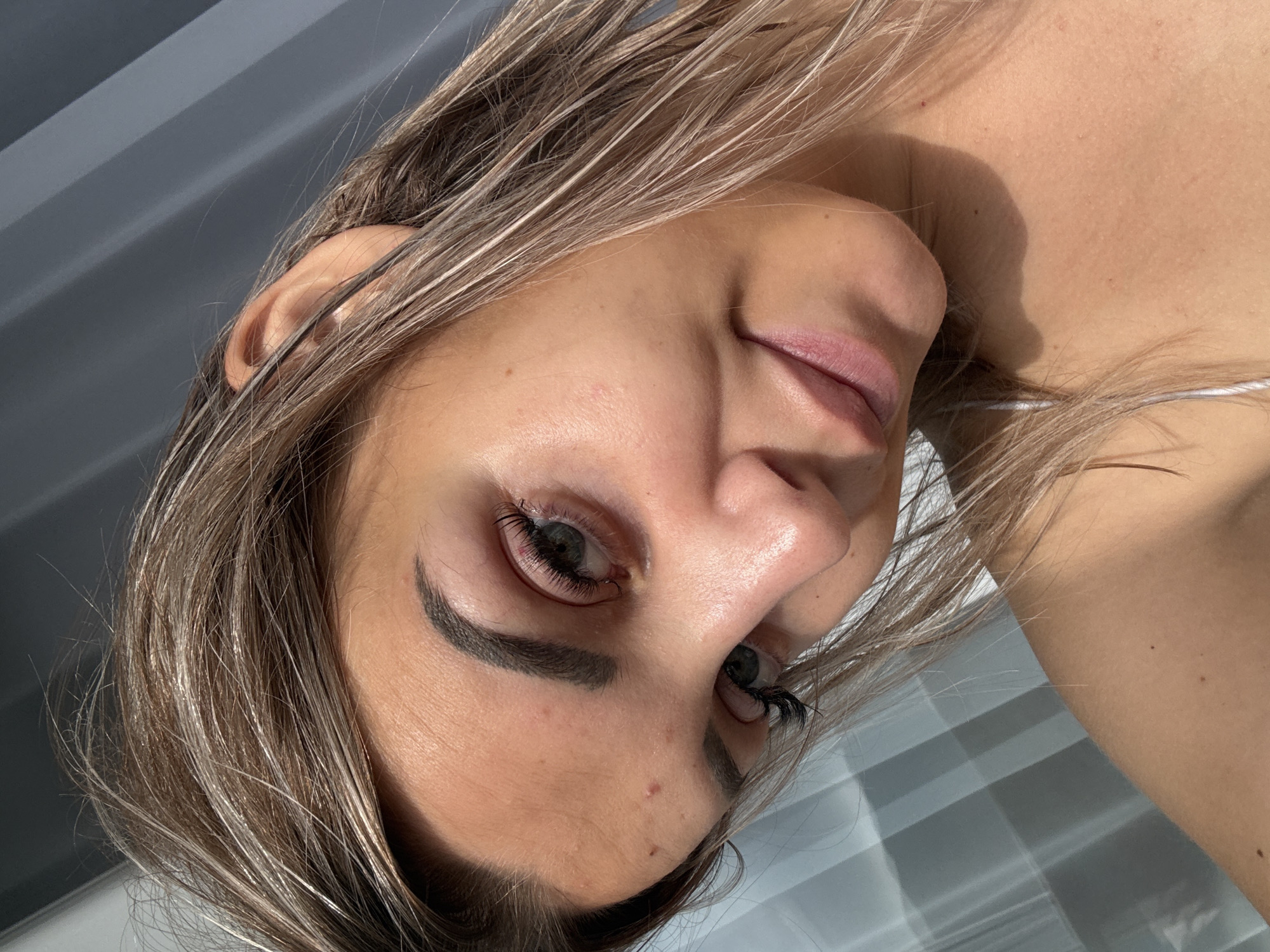 A close-up, low-angle, selfie-style shot of a young Caucasian woman. Her head is tilted to her left, with her face angled towards the camera. She has long, blonde-brown hair with lighter streaks, which is falling loosely around her face and shoulders. Her skin is lightly tanned and shows a few small blemishes. She is wearing subtle makeup, with defined eyebrows, mascara, and a pinkish lipstick. Her eyes are green. The background is blurred and consists of what appear to be vertical blinds or a window covering with horizontal light and shadow patterns. The lighting suggests it is daytime, with sunlight casting shadows and highlighting her features. The image does not contain any elements that would allow for the determination of its location in Kholmsk, Russia.