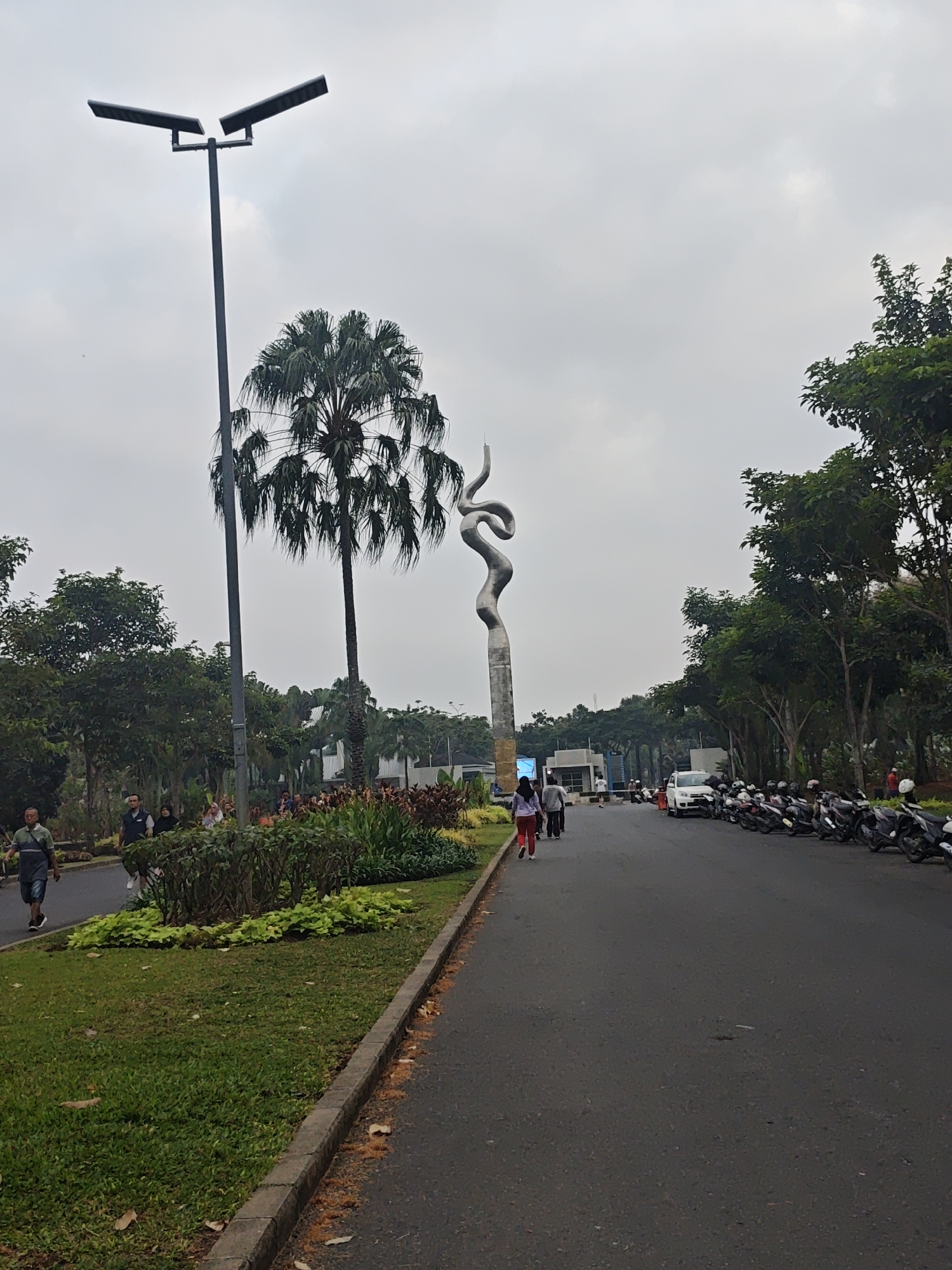 In Bacin, Indonesia, on a cloudy day, a paved road runs alongside a landscaped park. On the left, a man in a green shirt and shorts walks on a sidewalk bordering a grassy area with a curb. Further down the sidewalk, a group of people are walking, including a woman in red pants and a woman in a light-colored headscarf. Behind them is a large, distinctive sculpture resembling a coiled serpent or ribbon. A tall palm tree stands near the sculpture. On the right side of the road, a row of motorcycles are parked, and a white car is visible. Trees line the right side of the road, creating a verdant backdrop. A streetlamp with two angled lights stands tall on the left, near the palm tree. The environment appears to be a public space, possibly a park or entrance to a facility.