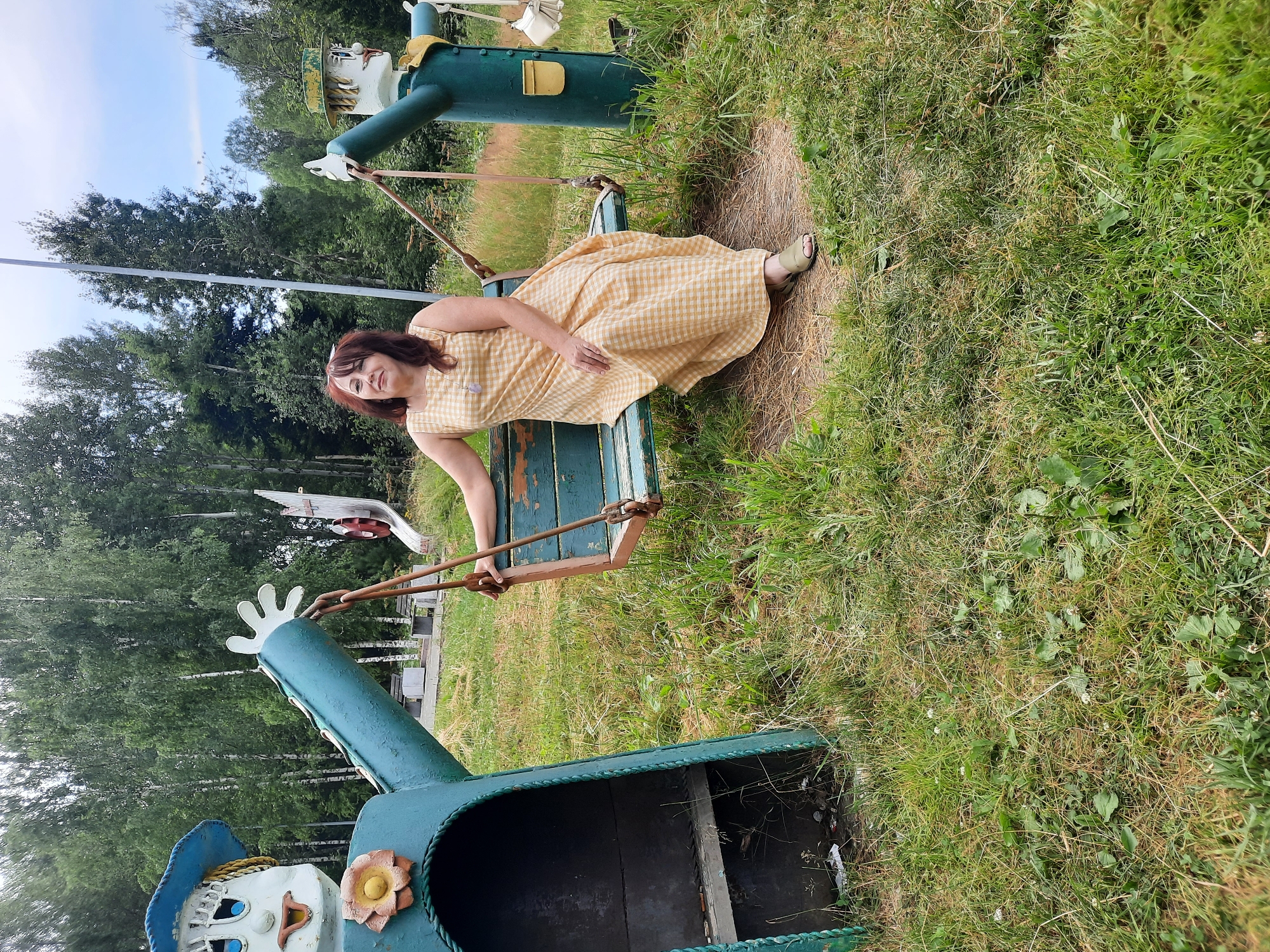 In Зеленогорский, Russia, a woman with dark hair, wearing a light yellow checkered dress and olive green sandals, sits on a weathered teal-colored swing. The swing is suspended by thick ropes connected to a tall, dark pole. To her left, a large, teal-colored scarecrow-like figure with a blue hat, painted face, and yellow flower accent stands beside the swing. To her right, another similar scarecrow figure, also teal with a hat and painted face, is visible. The figures appear to be part of a playground or decorative installation. The ground is covered in green grass with some dry patches and scattered small white flowers. In the background, a dense forest of green trees, primarily birches, creates a natural backdrop under a bright, partly cloudy sky.