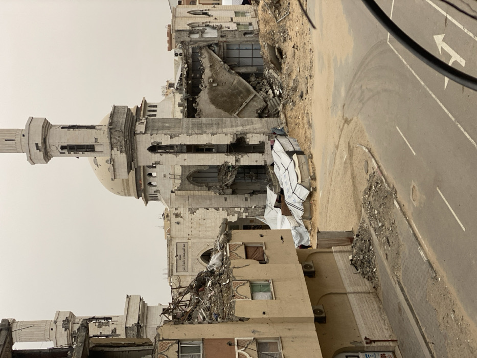 The image displays a severely damaged urban scene in Khan Yunis, Palestinian Territory, under an overcast sky. In the midground, a large mosque, featuring a central dome and two minarets, exhibits extensive structural failure. Its white-colored facade is heavily perforated, and large sections of walls are missing, revealing interior components and rebar. The right minaret is significantly fractured, with portions of its upper structure missing. Arabic script, specifically "مسجد المحمدية" (Al-Muhammadiyah Mosque), is visible above an arched entrance on the mosque's lower facade.
To the left, in the foreground, a multi-story, tan-colored building is also heavily damaged, with its upper levels collapsed into a large pile of rubble and exposed rebar. Windows are broken or missing, and small impact marks are visible on its exterior. Adjacent to this building, a paved asphalt road with white lane markings, including a turn arrow, curves across the bottom of the frame. A sidewalk runs parallel to the building, partially covered by dirt and small debris piles.
Between the foreground building and the mosque, on sandy, debris-strewn ground, several white tarp-covered structures resembling makeshift shelters are clustered. A single, small, indistinct figure in dark clothing is visible near these shelters, partially obscured by one of the tents. The overall environment is one of widespread structural devastation.