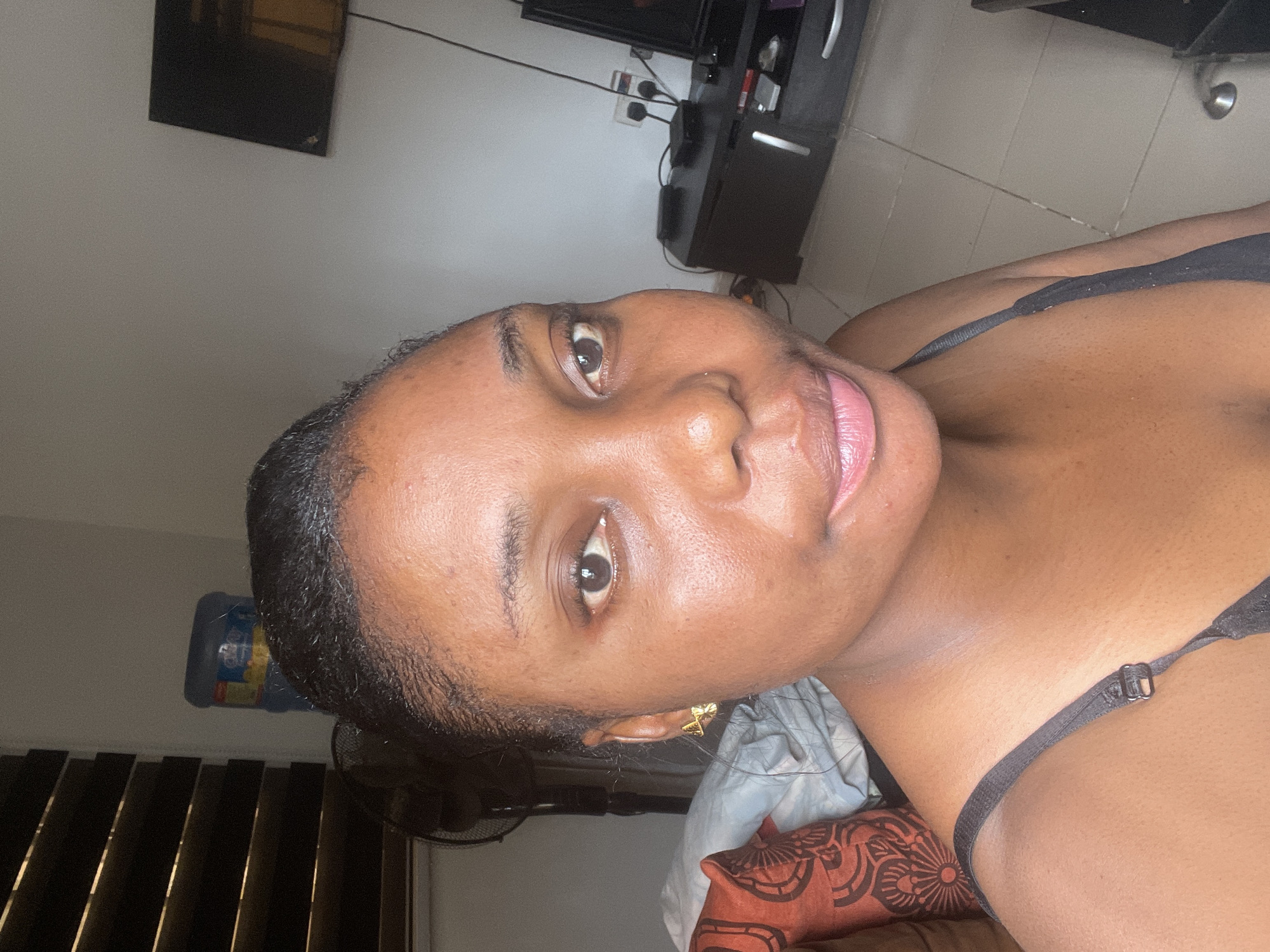 A close-up selfie depicts a dark-skinned woman with short, dark hair. She is wearing a black bra with adjustable straps. Her lips are pink and her eyes are brown. She appears to be indoors, lying on a bed with pillows. A television and entertainment center can be seen in the background. The precise location in Abuja, Nigeria, cannot be determined from the image.
