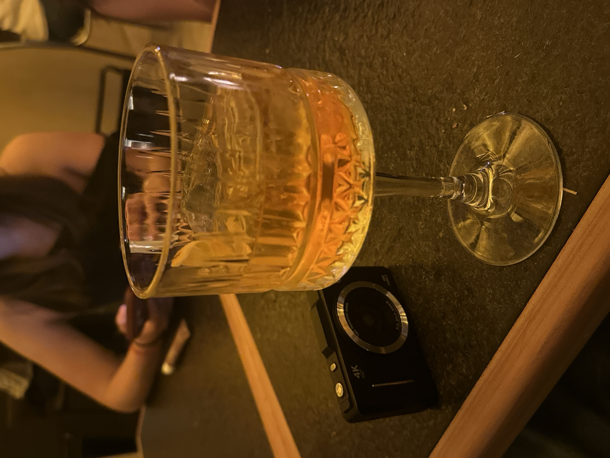 The image shows a glass of amber liquid, likely a cocktail, resting on a dark, possibly granite countertop. Next to the glass is a small black digital camera labeled "4K". The countertop appears to be part of a table in a dimly lit indoor setting. In the background, a person is seated, visible by their arm and shoulder. Given the context of the prompt requesting the location of San Andrés Cholula, Mexico, it is plausible that the photo was taken in a bar or restaurant in this city, but specific details to verify the location (e.g. logos, names) are absent.