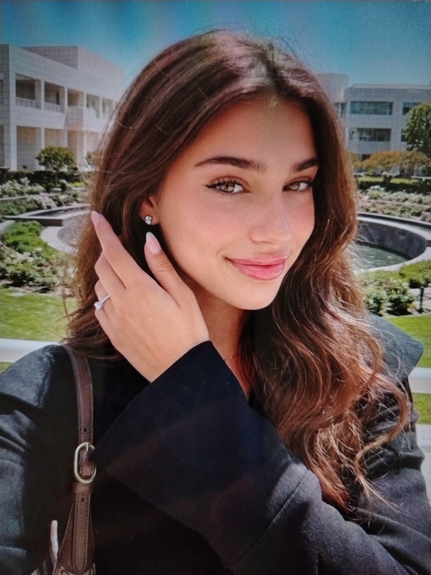This is a head and shoulder shot of a young woman with long, wavy brown hair and brown eyes. She is wearing a black jacket, and a brown purse strap is visible on her left shoulder. She has a diamond stud earring in her left ear and a diamond ring on her left ring finger. Her left hand is raised to her head. In the background, there is a modern building with white columns and a circular fountain with greenery. The sky is a clear blue. The location is not identifiable as Stavropol, Russia.