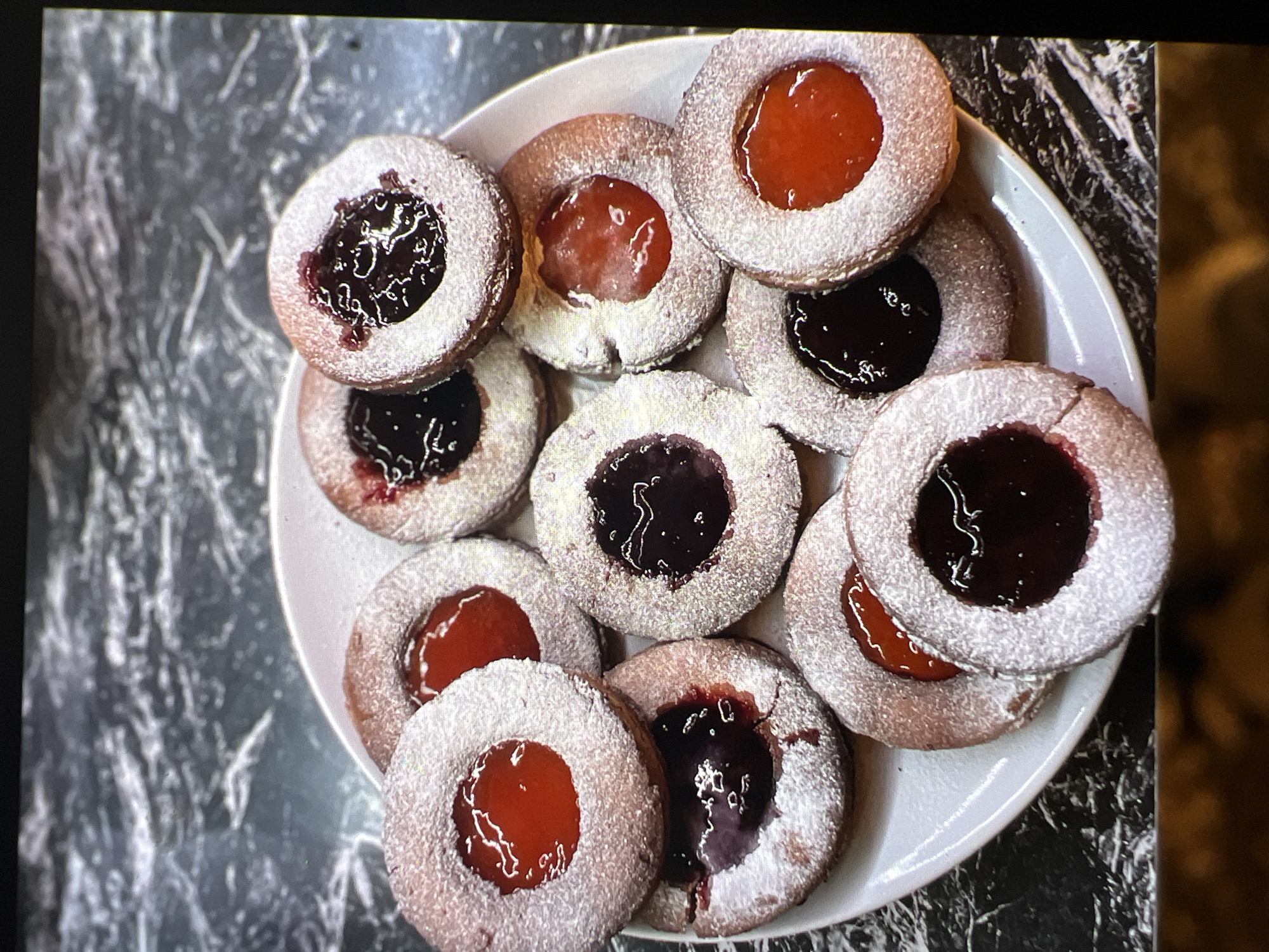 A plate of jam-filled cookies is arranged on a white plate, which is placed on a dark, marbled surface. The cookies are round with a thumbprint-like indentation in the center, filled with either red or dark purple jam. They are dusted with powdered sugar. The background is out of focus, showing the marbled pattern. There are no people or actions depicted in the image. The scene appears to be a still life photograph.