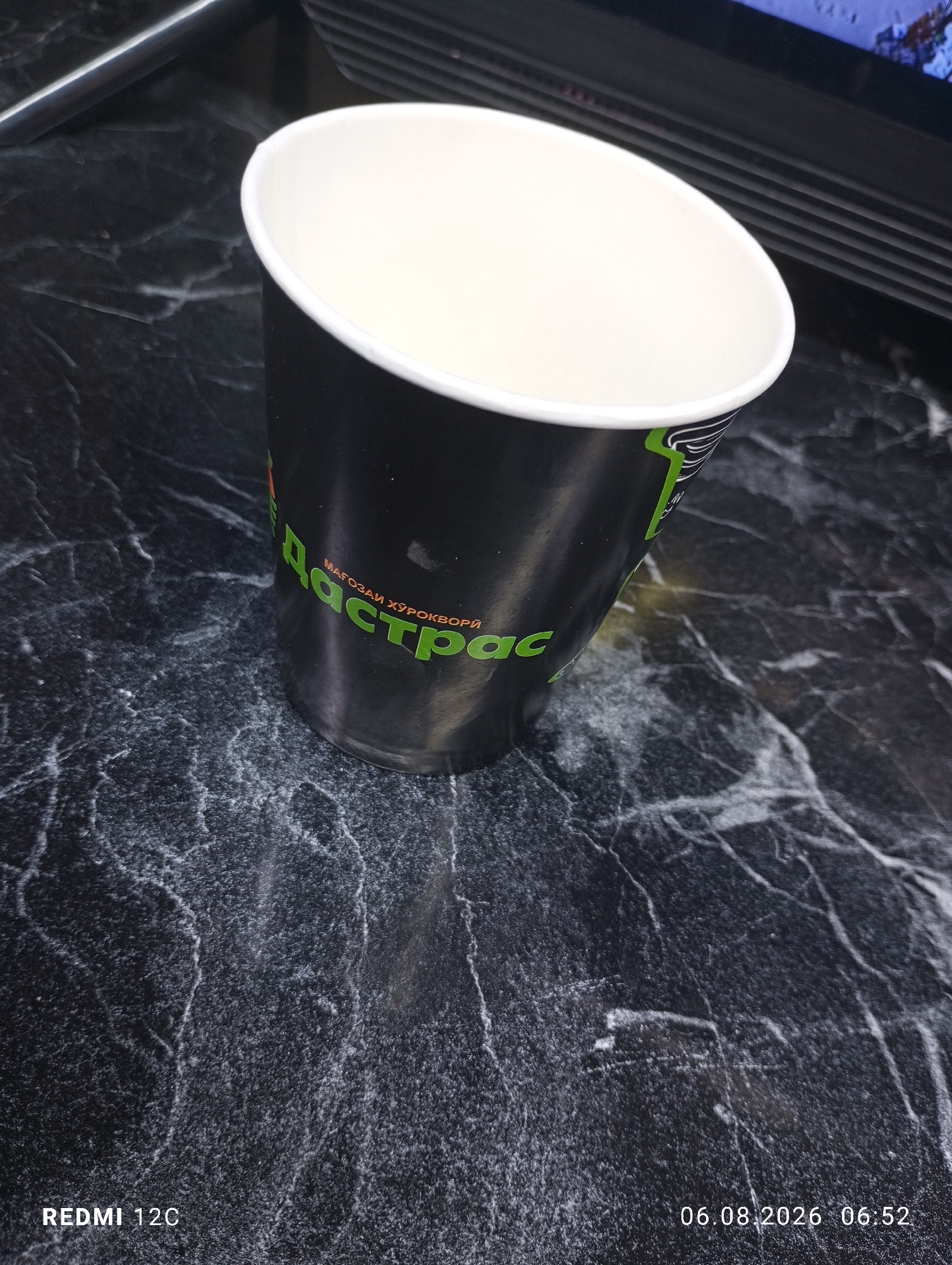 An overhead shot shows a black paper cup with a white interior and a green logo on a black marble-patterned surface. The logo on the cup reads "Даст рас" in large green letters, with smaller text above it. To the left of the cup, the text "REDMI 12C" is visible, indicating the device used to take the photo. In the lower right corner, a date and time stamp reads "06.08.2026 06:52". The background shows a portion of a dark, possibly electronic device. The scene is devoid of any individuals or specific environmental details that would indicate a location in Buston, Tajikistan, or any other specific city. The lighting appears to be artificial and directed from above.